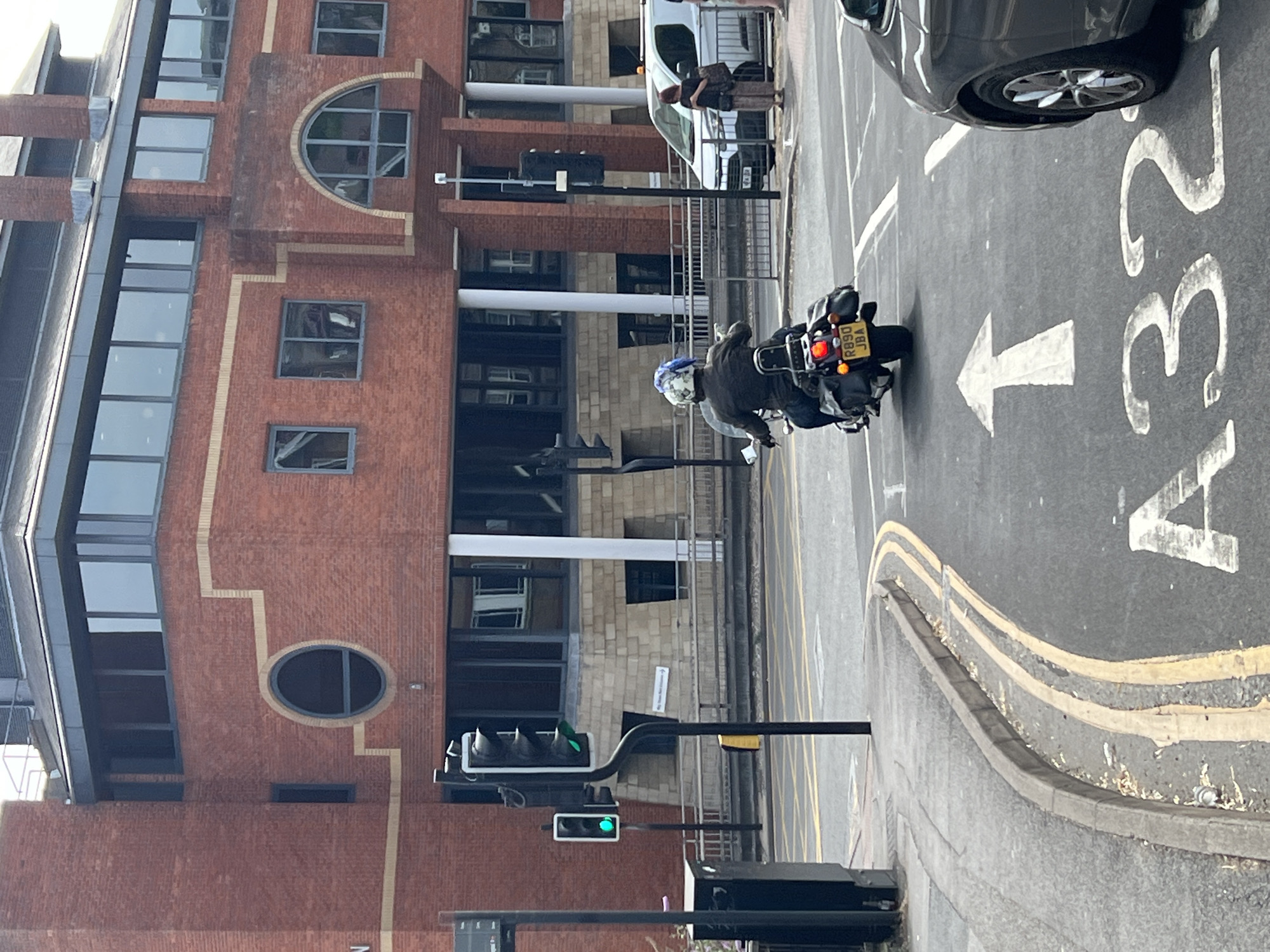 The image shows a street scene in Guildford, United Kingdom. A motorcycle with a license plate that reads "R890 JBA" is in the foreground, heading away from the viewer. The motorcycle has a rider wearing a helmet with a blue and white pattern. Behind the motorcycle, a dark-colored car is parked on the right side of the road. In the background, there is a large brick building with multiple windows and architectural details. Traffic lights are visible on the left side of the image, with a green light illuminated. The road has lane markings and a sign with the text "A32" stenciled on it. There is a pedestrian crossing in the distance. The weather appears to be overcast.