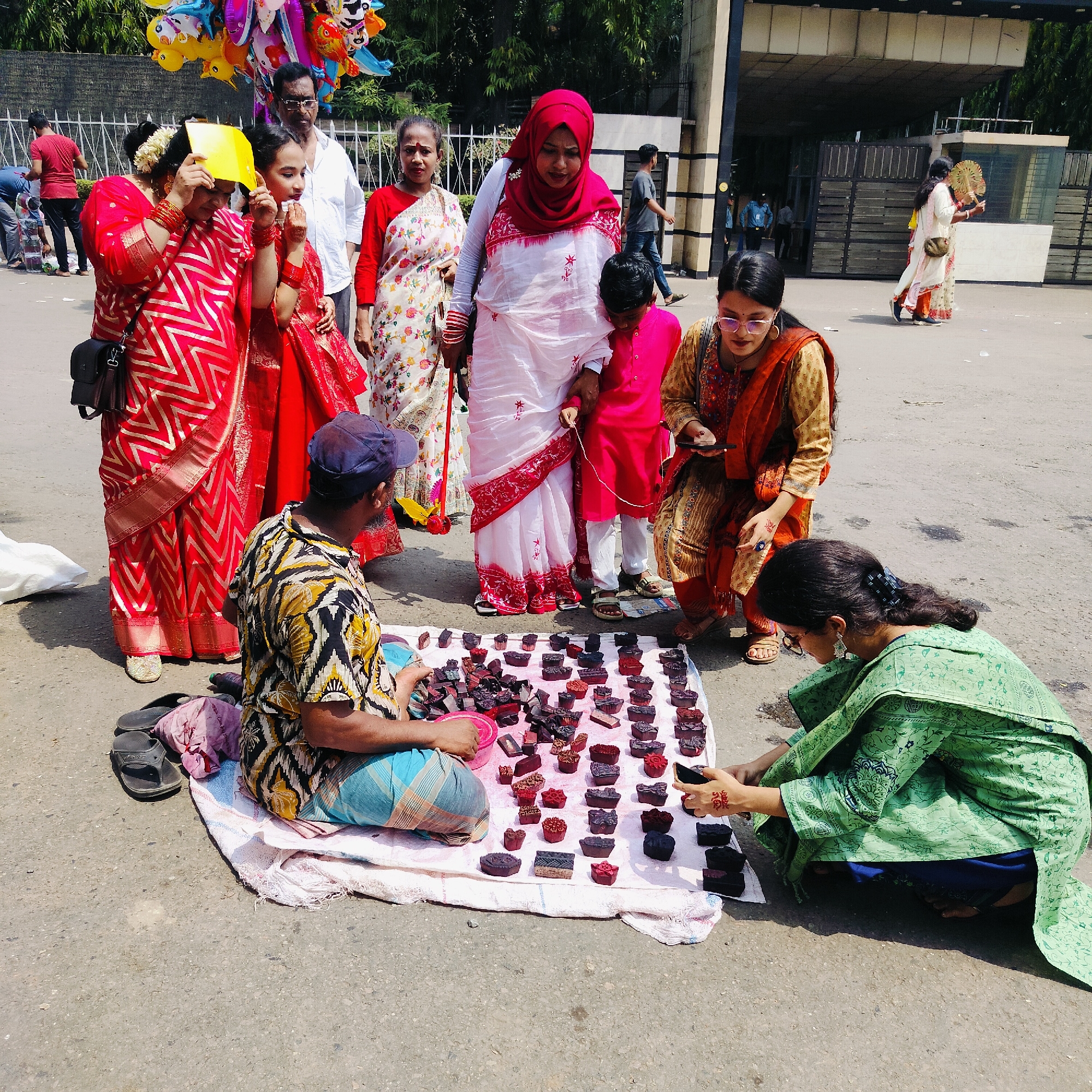The image depicts an outdoor scene, likely a street or public space in Dhaka, Bangladesh, based on the clothing and general ambiance. A vendor, wearing a patterned shirt and blue cap, is seated on the ground behind a white cloth displaying numerous small, dark-colored stamps or blocks. Two women are examining the stamps. One woman, kneeling and wearing a green outfit, is looking at the stamps on the cloth. The second, kneeling and wearing a yellow and orange outfit, appears to be taking a picture with her phone. Several other people are standing in the background. The people in the background are all wearing traditional Bengali clothing. To the right of the group, two women are holding hand fans. A large building facade is in the background, suggesting a government facility or public building. The overall impression is that of a lively street scene in Dhaka.