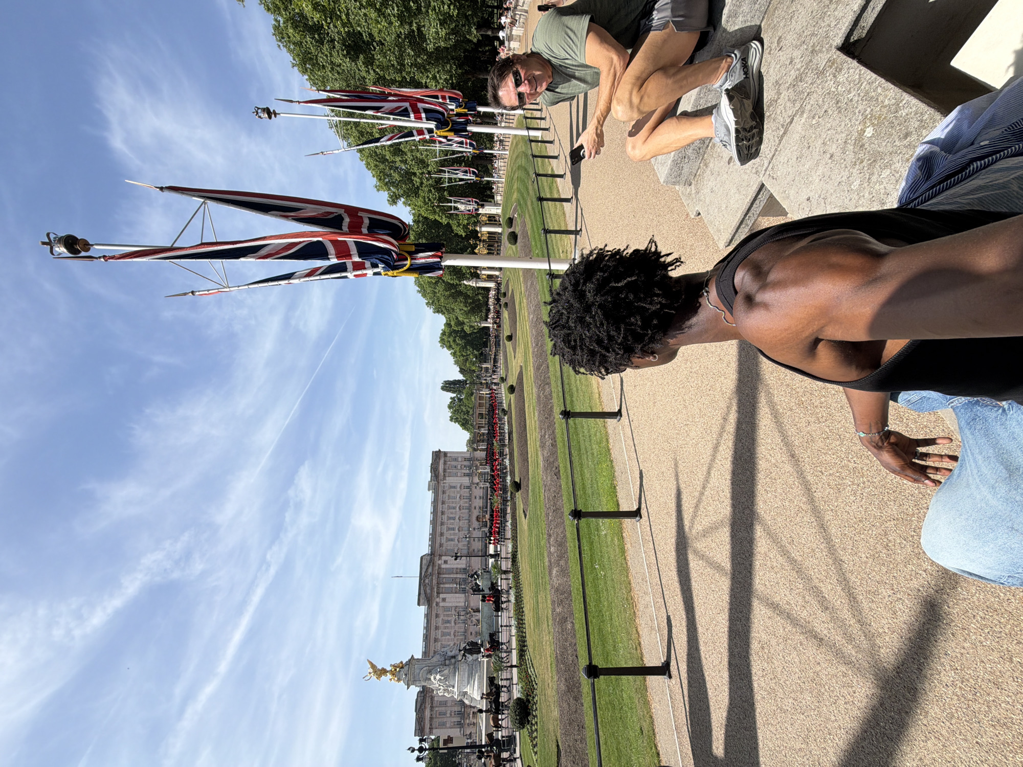 The image captures a scene near Buckingham Palace in London, United Kingdom. The foreground shows two people on a paved surface, with one person wearing a black tank top and light-colored denim jeans sitting near the edge and another person in a green shirt and dark shorts squatting. In the background, Buckingham Palace stands prominently with a line of red-uniformed guards visible. Union Jack flags are displayed on flagpoles, and a sunny sky with light clouds is overhead.