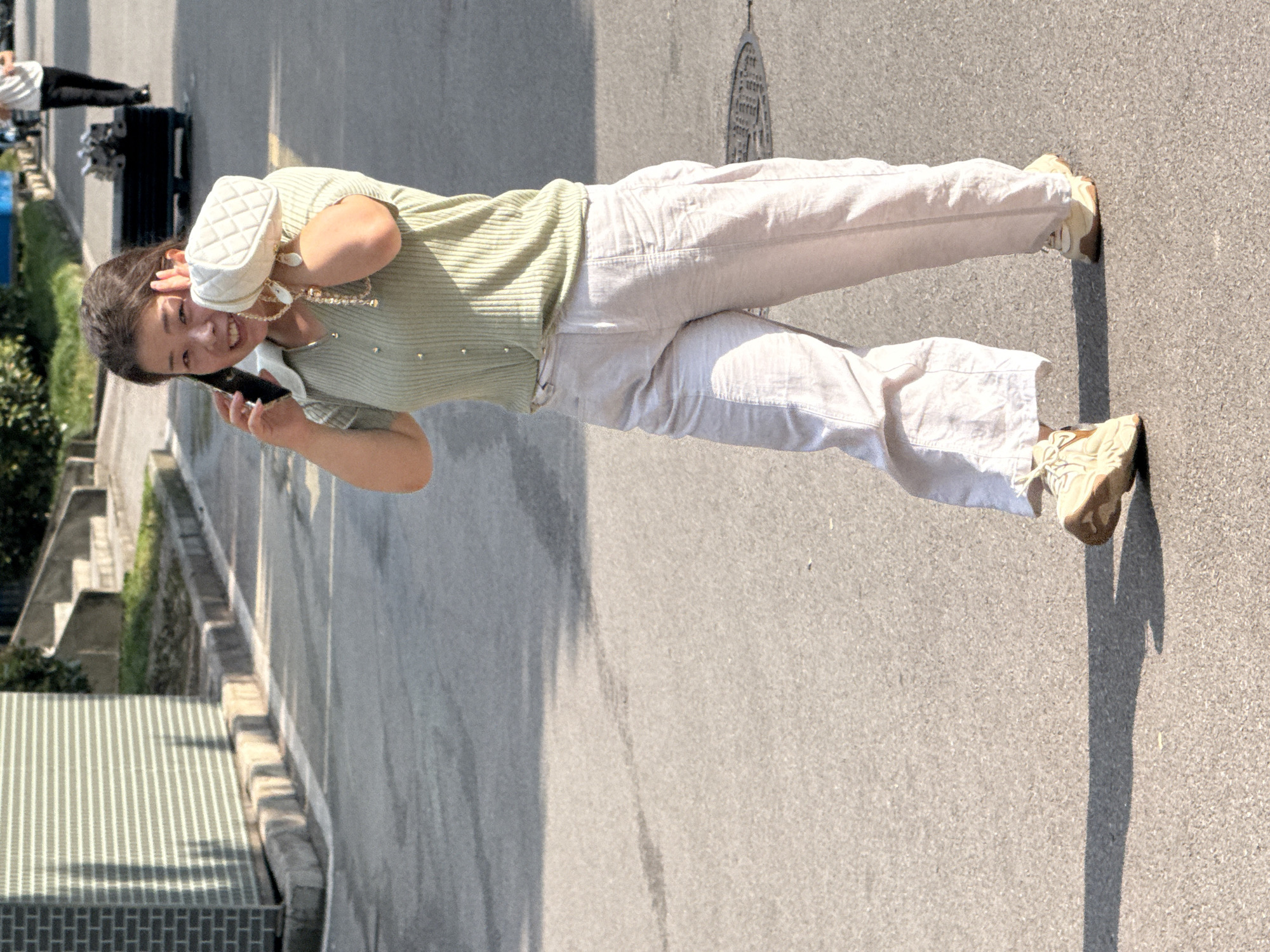 A young woman stands on an asphalt surface, talking on her smartphone. She is wearing a light green, ribbed short-sleeved shirt with small pearls on the front, and loose, cream-colored pants. Her feet are adorned with light tan sneakers. She holds a small, quilted white purse in her left hand, which is near her face, and her right hand is holding the phone to her ear. Her expression appears cheerful and engaged in conversation.
The scene is outdoors on a sunny day, indicated by the strong shadows cast on the ground. The background includes a concrete wall or building to the right, and a tiled structure and some greenery to the left, suggesting an urban or semi-urban environment. The orientation of the image is vertical, with the woman appearing to be walking or standing in a space that suggests a street or walkway. The context provided places this scene in Jiangjin, China. No visible text is present in the image.