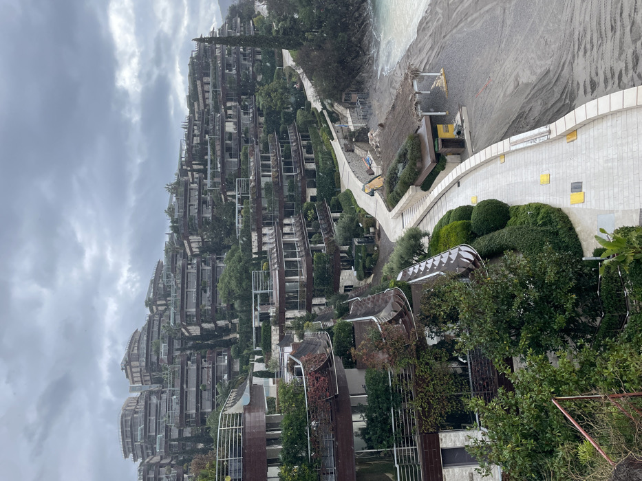 The image presents an elevated view of a terraced resort complex on a hillside in Budva, Montenegro, under a heavily overcast sky. The foreground and midground feature numerous modern, multi-story buildings constructed into the slope, characterized by their light-colored stone or concrete facades, extensive dark-brown balconies, and abundant green vegetation on rooftops and terraces. White metal railings are visible on balconies and structural edges.
A light-colored, paved walkway, possibly stone or concrete, winds horizontally through the midground, following the contour of the coastline. This pathway is lined with meticulously maintained green landscaping, including shrubs and trees. Several yellow square markers are embedded in the pathway's surface. Adjacent to the pathway, on a dark grey-brown beach, a structure with a thatched roof and open supports is visible, accompanied by a smaller, enclosed utility-style building featuring yellow doors or panels. The beach exhibits disturbed sand or pebbles, possibly from recent grooming or vehicle activity. The Adriatic Sea, with light blue-green water and gentle waves, borders the beach on the right side of the frame, receding towards a distant coastline. No individuals are present within the scene. A red-colored metal barrier is partially visible in the lower-left corner.