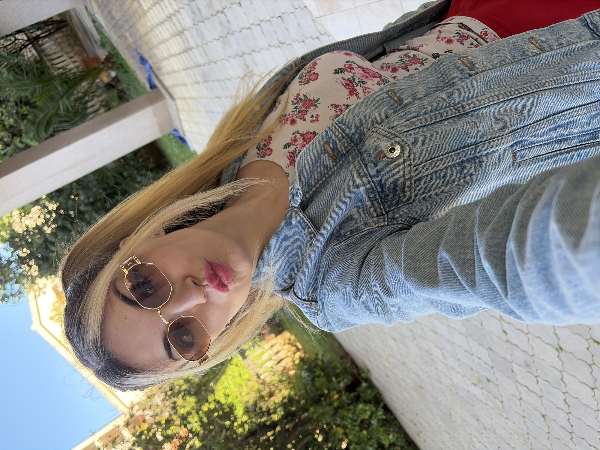 A woman is posing for a selfie. She is wearing sunglasses, a light denim jacket, and a floral shirt. Her hair is blonde. The background shows trees and a light-colored tiled surface. The location appears to be outdoors, possibly a patio or garden in Budva, Montenegro, based on the light, foliage, and architectural style.
