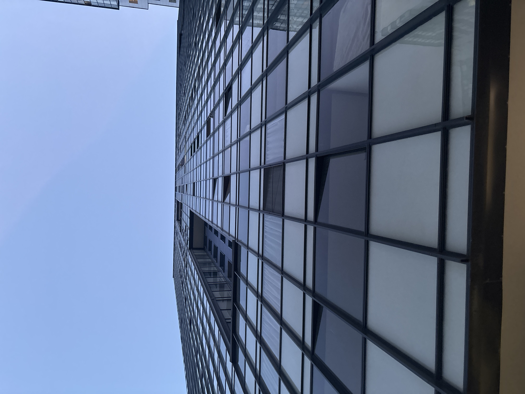 This image is a low-angle, full shot of a modern skyscraper with a glass facade under a clear blue sky. The building's architecture is characterized by a grid of dark-framed windows, some of which are open. The perspective emphasizes the height and scale of the structure, as it recedes upwards towards the sky. The glass reflects the sky and surrounding environment, creating a sleek and contemporary aesthetic. The lighting suggests it is daytime, with soft shadows and a bright sky. The scene is devoid of people or any discernible activity, focusing solely on the architectural form of the building.