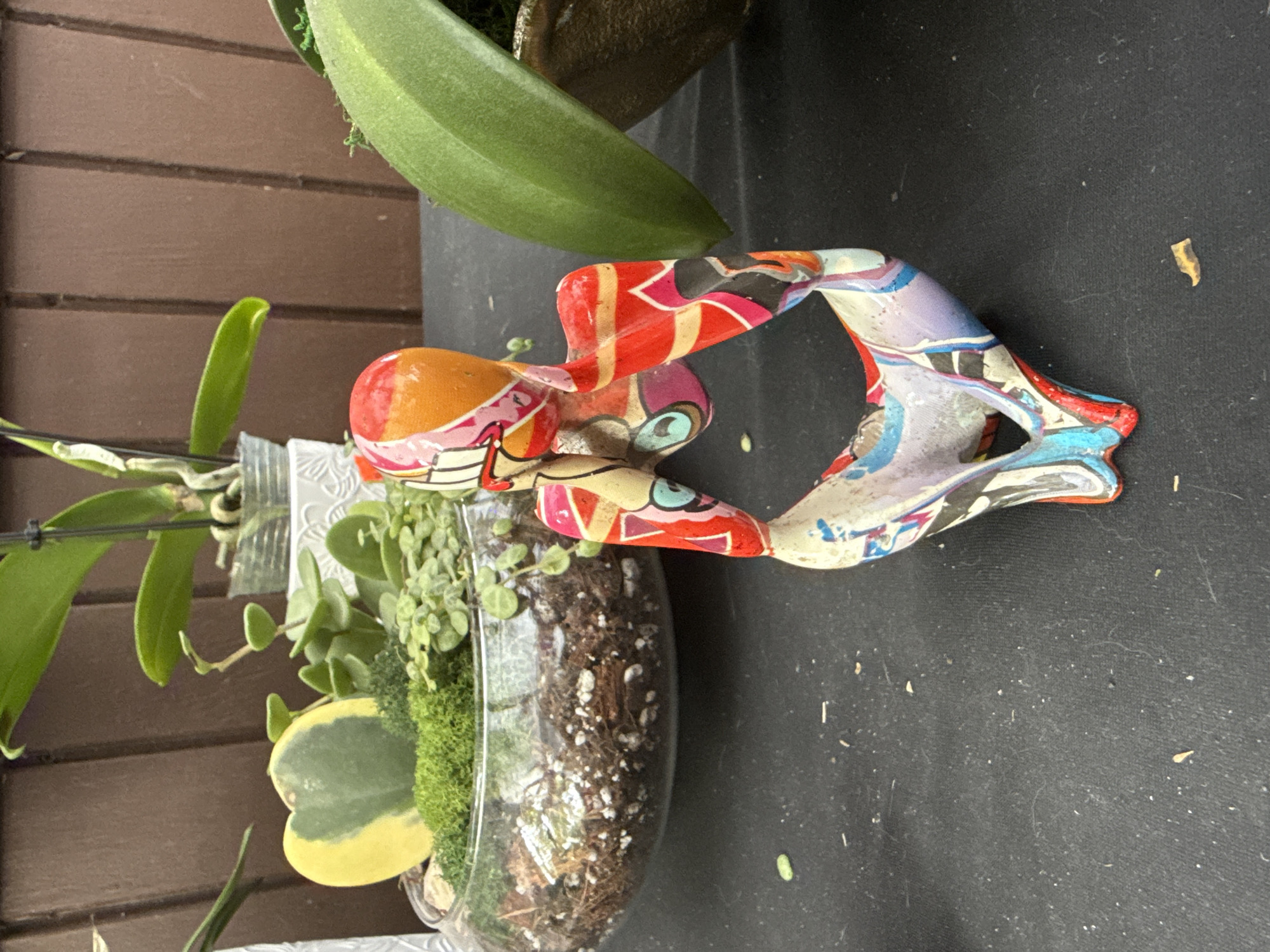 The image shows a still life scene composed of a small, clear glass bowl containing a plant arrangement, alongside a colorful, patterned figurine, set against a backdrop of brown horizontal wooden planks. The plant arrangement features a variety of greenery, including a heart-shaped variegated Hoya leaf, moss, and other trailing plants set in soil. The figurine, predominantly orange, red, and blue, appears to be an abstract sculpture. The surface beneath the arrangement and figurine is a dark, possibly black material. The location of the scene is Davie, United States of America.