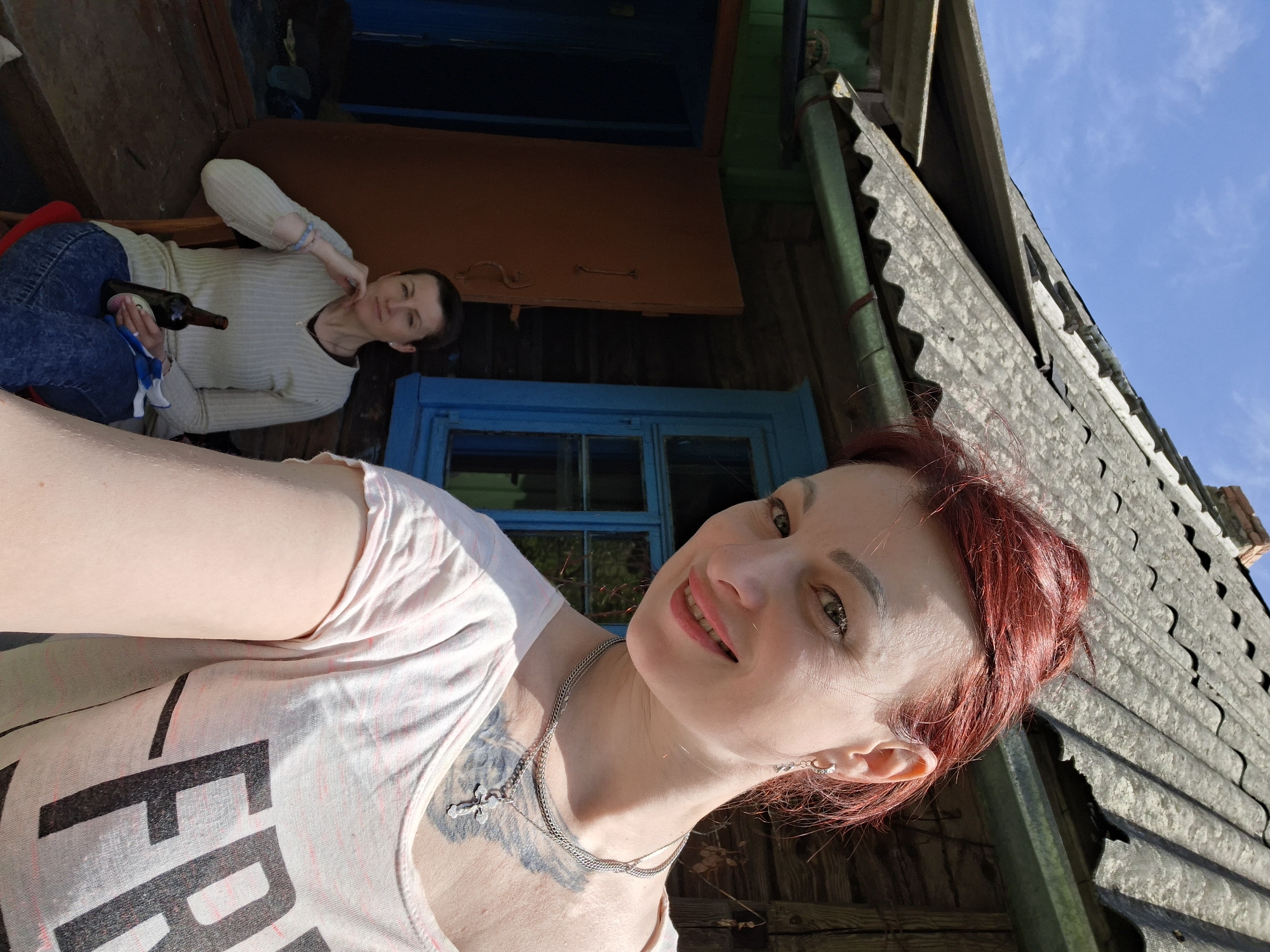 The image appears to be a selfie taken outdoors in Сваткаўскі сельскі Савет, Belarus. The foreground features a woman with red hair, wearing a light pink t-shirt with a large black graphic and a silver necklace. She is smiling at the camera. Her right ear has an earring.
Behind her and slightly to the left, another woman is reclining in a chair, holding a bottle of wine. She is wearing a light-colored sweater and blue jeans.
The backdrop consists of a wooden building with blue window frames and a corrugated metal roof. The sky is visible in the upper right corner, indicating it's a sunny day. The building appears to be old.