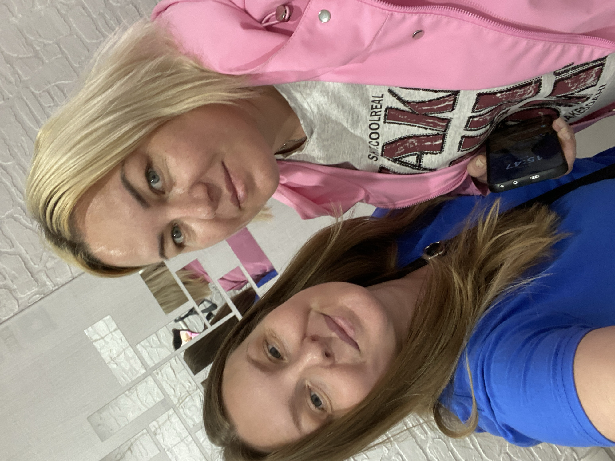 This image captures two adult females in a close-up, selfie-style photograph indoors, located in the city of Noviy, Russia. The woman on the left, positioned in the foreground, has long, light brown hair and light-colored eyes, and is wearing a royal blue short-sleeved top. She presents a subtle smile towards the camera. The woman on the right has shoulder-length blonde hair and light-colored eyes, and is wearing a pink puffer jacket over a light grey t-shirt featuring distressed dark red lettering that reads "SK. COOL REAL" followed by partially visible text. She holds a dark-colored smartphone in her left hand, which displays the time "15:47". Both individuals appear to be in their 30s or 40s. The background consists of a light-colored, textured wall with multiple irregularly arranged rectangular mirror tiles reflecting fragmented portions of the subjects and their attire.