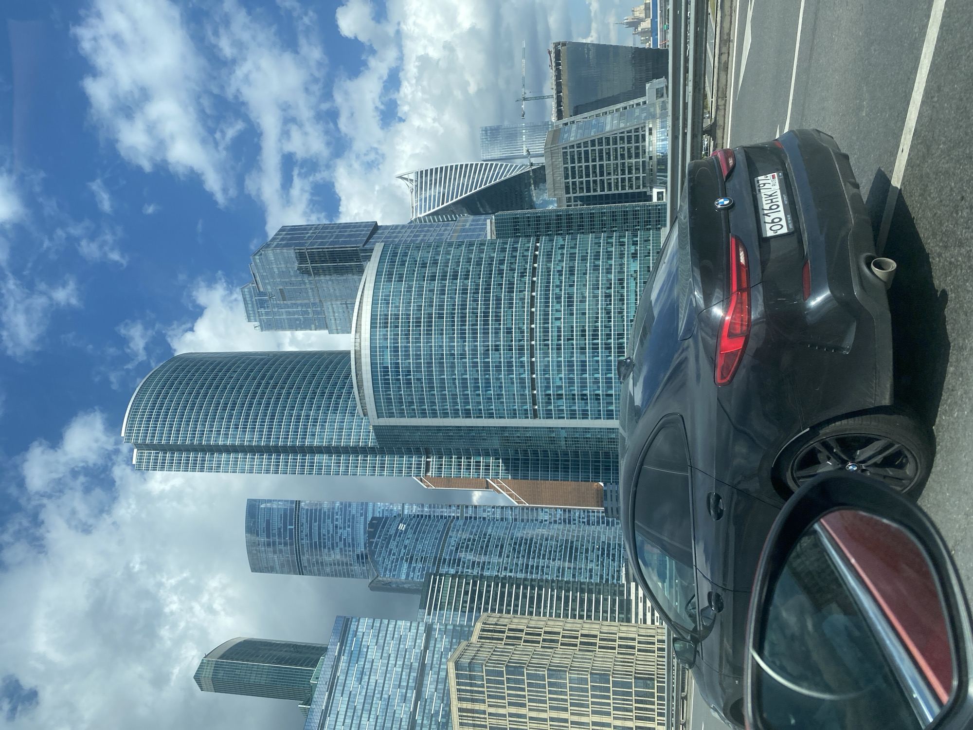 The image captures a view of the Moscow International Business Center, commonly known as Moscow-City, in Moscow, Russia, from a moving vehicle. In the foreground, the rear of a black BMW sedan is visible, its license plate reading "0616HK 795 RUS". The car is on an elevated road, indicated by the guardrail and road markings. To the left of the BMW, the side mirror of another vehicle reflects the environment, showing part of its interior and the road ahead.
The background is dominated by the modern skyscrapers of Moscow-City, characterized by their glass and steel facades. Prominent buildings include the curved residential complex on the left, and various office towers with distinct architectural designs, such as the twisted building in the center-right. The sky is partly cloudy, with patches of blue sky visible between the white clouds, suggesting a bright but dynamic weather condition. Construction cranes are also visible in the distance, indicating ongoing development in the area. The overall scene depicts a typical urban landscape of a modern metropolis, with contemporary architecture and infrastructure.
