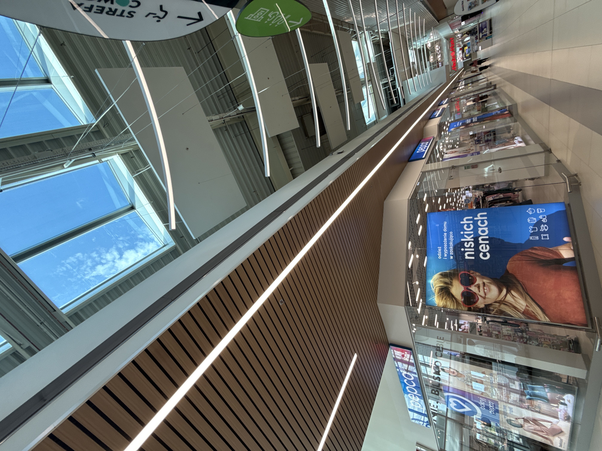 The image captures an interior view of a modern shopping mall in Bielany Wrocławskie, Poland. The ceiling features a large skylight allowing natural light to filter into the spacious atrium, illuminating the contemporary architecture. Linear LED lights are strategically placed along the ceiling and decorative wooden panels, creating a warm and inviting atmosphere.
In the foreground, a shop with a glass facade displays various products. A large advertisement on the right features a young woman with blonde hair and red heart-shaped sunglasses, promoting "niskich cenach" (low prices). The Pepco store logo is visible on the left. Other storefronts and signage are visible further down the mall, suggesting a variety of retail establishments.
The flooring is a combination of light-colored tiles and darker gray strips, creating a patterned walkway. Several individuals are present in the mall, walking in the distance, indicating it is open for business. The overall impression is one of a bright, clean, and contemporary retail environment.