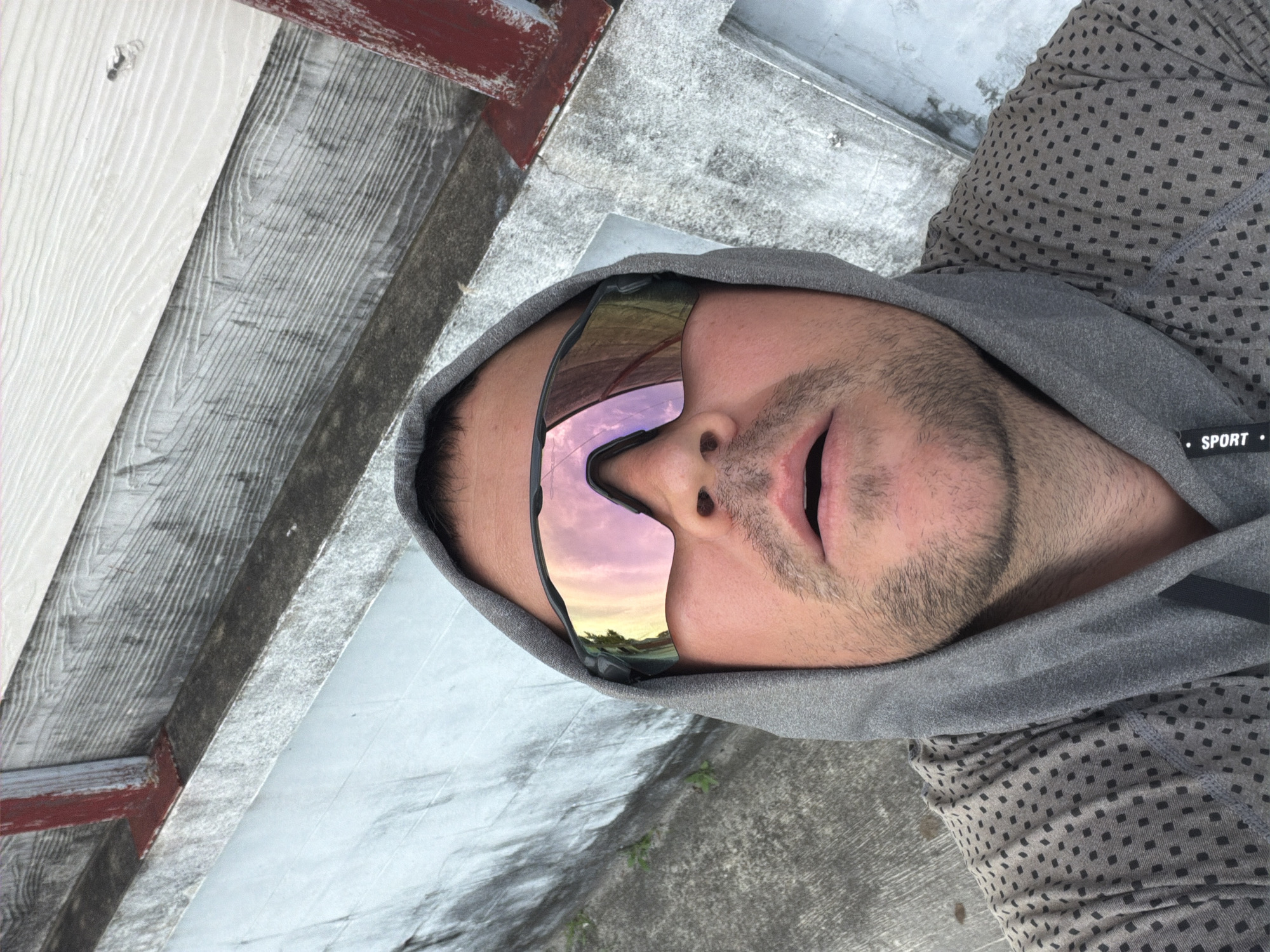 A man wearing a gray hooded sweatshirt and sunglasses takes a selfie outdoors. The sunglasses reflect a pinkish sky with clouds and trees. He has stubble on his face and his mouth is slightly open. The background consists of concrete and weathered wood. The man's hood is up, partially obscuring his head. The scene appears to be in Baan Mae Nam, Thailand, based on the available information.