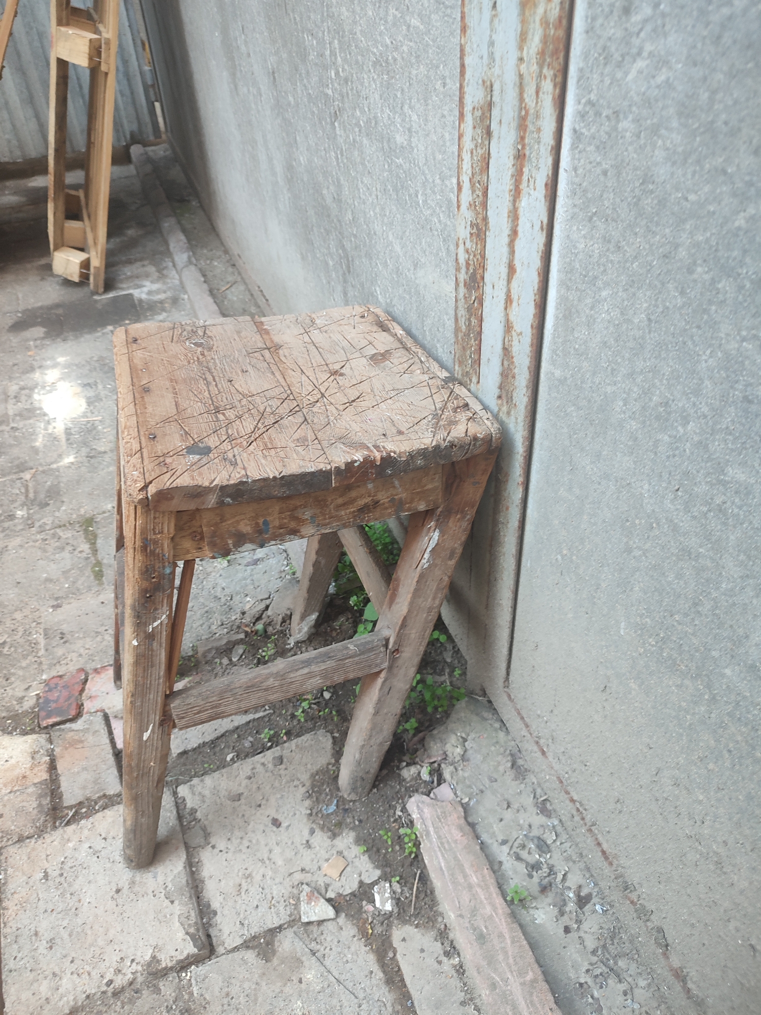 A rustic, weathered wooden stool is positioned at an angle against a textured, gray concrete wall. The stool has a square seat marked with numerous scratches and indentations, indicative of heavy use. Its legs are constructed from rough-hewn wooden beams, braced by additional wooden supports. The stool is situated on a cracked, uneven concrete ground, interspersed with patches of dirt and small green plants. To the left of the stool, a portion of a wooden structure, possibly part of a larger shelving unit or workbench, is visible. A corrugated metal element, showing signs of rust, runs vertically along the wall behind the stool, suggesting an outdoor or industrial setting. The overall impression is one of an unkempt, functional area, possibly in a backyard, workshop, or alleyway in Chernivtsi, Ukraine. No people are present in the image.
