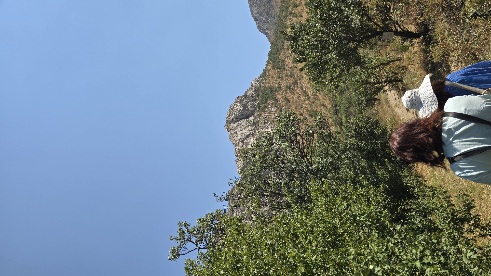 In the city of Terekty, Kazakhstan, two individuals are walking away from the camera on a dirt path that winds through a mountainous, arid landscape. The sky above is clear and bright blue. In the foreground, lush green foliage borders the path on the left side. The two people are dressed casually, with one wearing a light blue t-shirt and a backpack, and the other wearing a wide-brimmed white hat and a blue garment. Ahead of them, the path leads towards a rocky, barren mountain peak. The terrain is characterized by sparse vegetation, with shrubs and dry grasses scattered across the hillsides. The overall scene suggests a hike or exploration in a natural, outdoor environment.