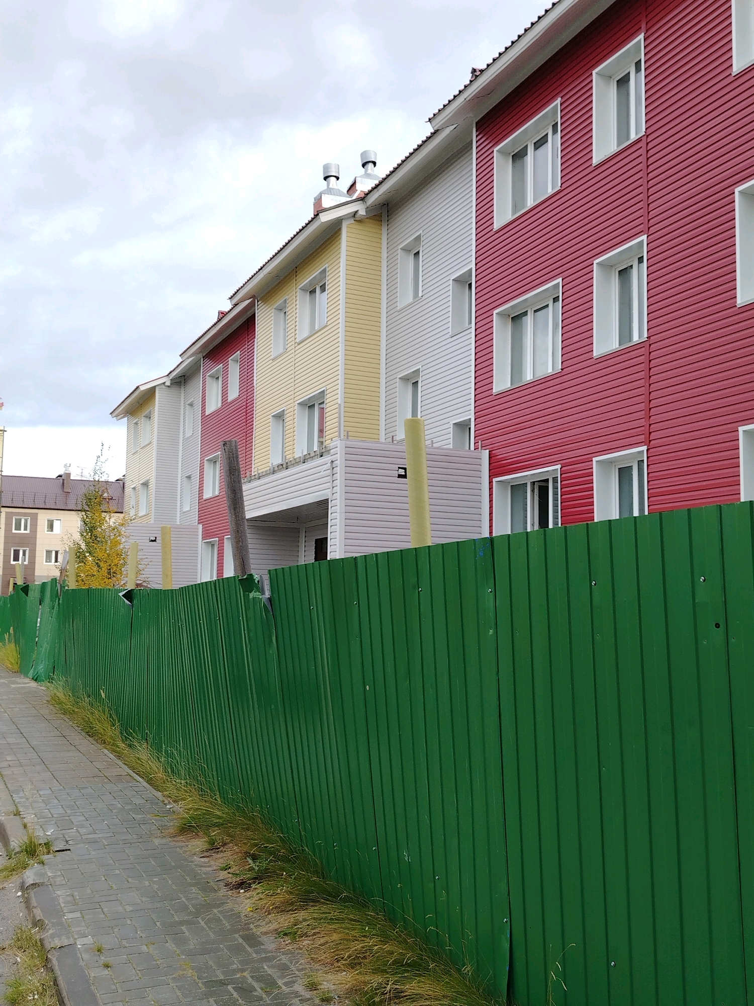 A row of multi-story apartment buildings in Muravlenko, Russia, are visible behind a tall, green corrugated metal fence. The buildings are painted in distinct sections of red, white, and yellow, with white-framed windows. The sky is overcast with gray clouds. A paved walkway runs alongside the fence, with patches of dry grass growing between the paving stones and along the base of the fence. The scene appears to be a residential area.