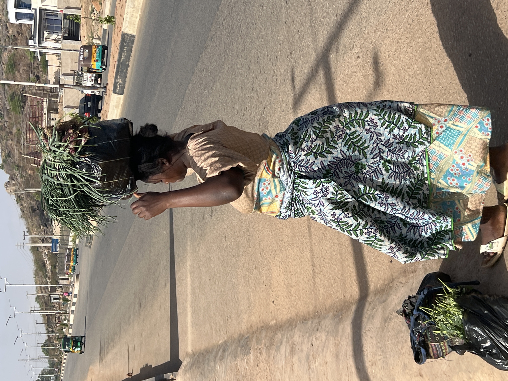 The image captures a woman walking on a wide, paved road in Jos, Nigeria, under bright sunlight. She is carrying a large, black-bagged bundle of green leafy vegetables, resembling green onions or scallions, balanced on her head. Her right arm is bent at the elbow, with her hand held in front of her, possibly for balance or to adjust her load. She wears a light brown, short-sleeved top and a patterned skirt or wrapper with green, blue, and brown floral and geometric designs. She is wearing sandals.
Next to her, on the ground, another black bag filled with similar green vegetables is visible. The road appears to be a main street or highway, with a black and white striped curb dividing it. Long, distinct shadows are cast on the road, indicating the sun is high in the sky, likely mid-morning or late afternoon.
In the background, several buildings line the road, and arid, rocky hills are visible under a clear sky. Various vehicles, including auto-rickshaws (locally known as Keke Napep) and cars, are present on the road further down. One yellow and green auto-rickshaw has the text "JIR-19-J3" visible on its back. Street lights or utility poles are also spaced along the roadside. The scene depicts a moment of daily life, likely a vendor transporting produce in a bustling, sunny environment.