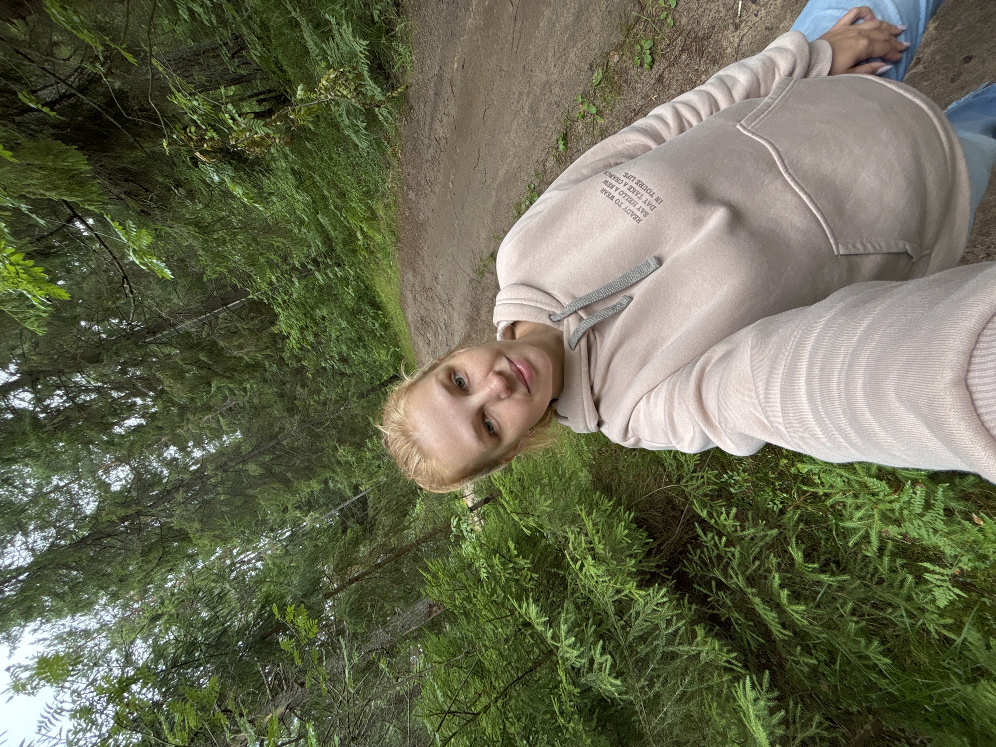 A young woman with blonde hair tied back in a bun is taking a selfie in a forest. She is wearing a light pink hooded sweatshirt and blue jeans. The forest is dense with tall evergreen trees and lush green undergrowth. A dirt path winds through the trees behind her. The woman is smiling slightly at the camera. The location is identified as Anteplevo (Okhotinskoe S/P), Russia.