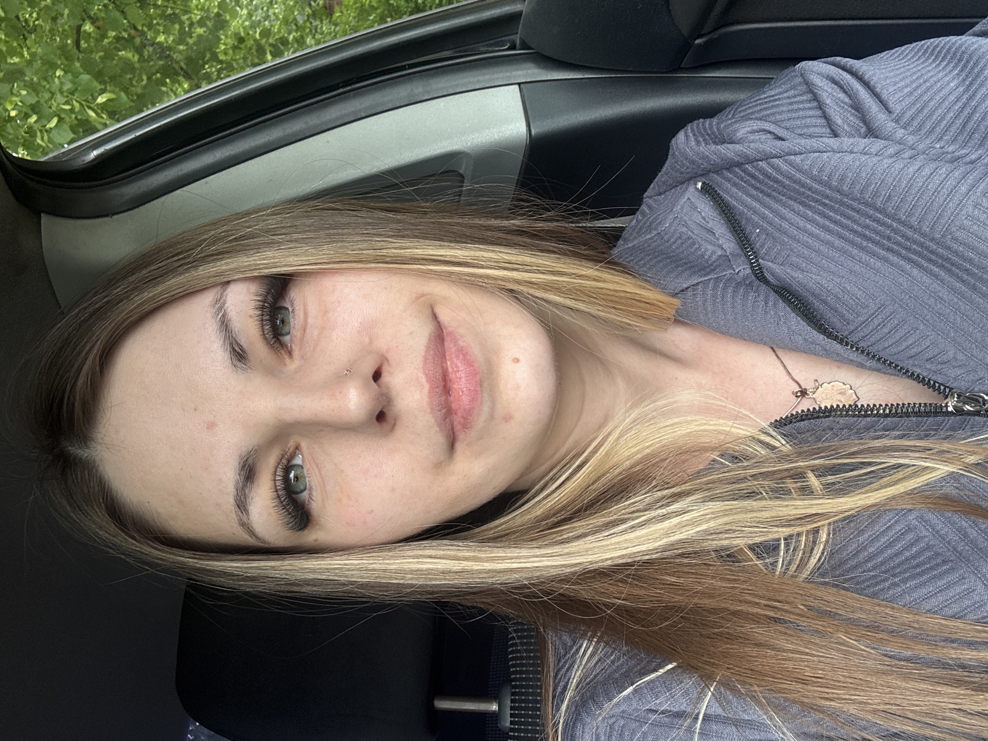A blonde-haired woman with green eyes is seated in a vehicle. She has a nose piercing and is wearing a gray zip-up shirt. The car's interior is visible, and the background shows foliage outside the window. The image does not contain information to identify Shevlyakovo, Russia.