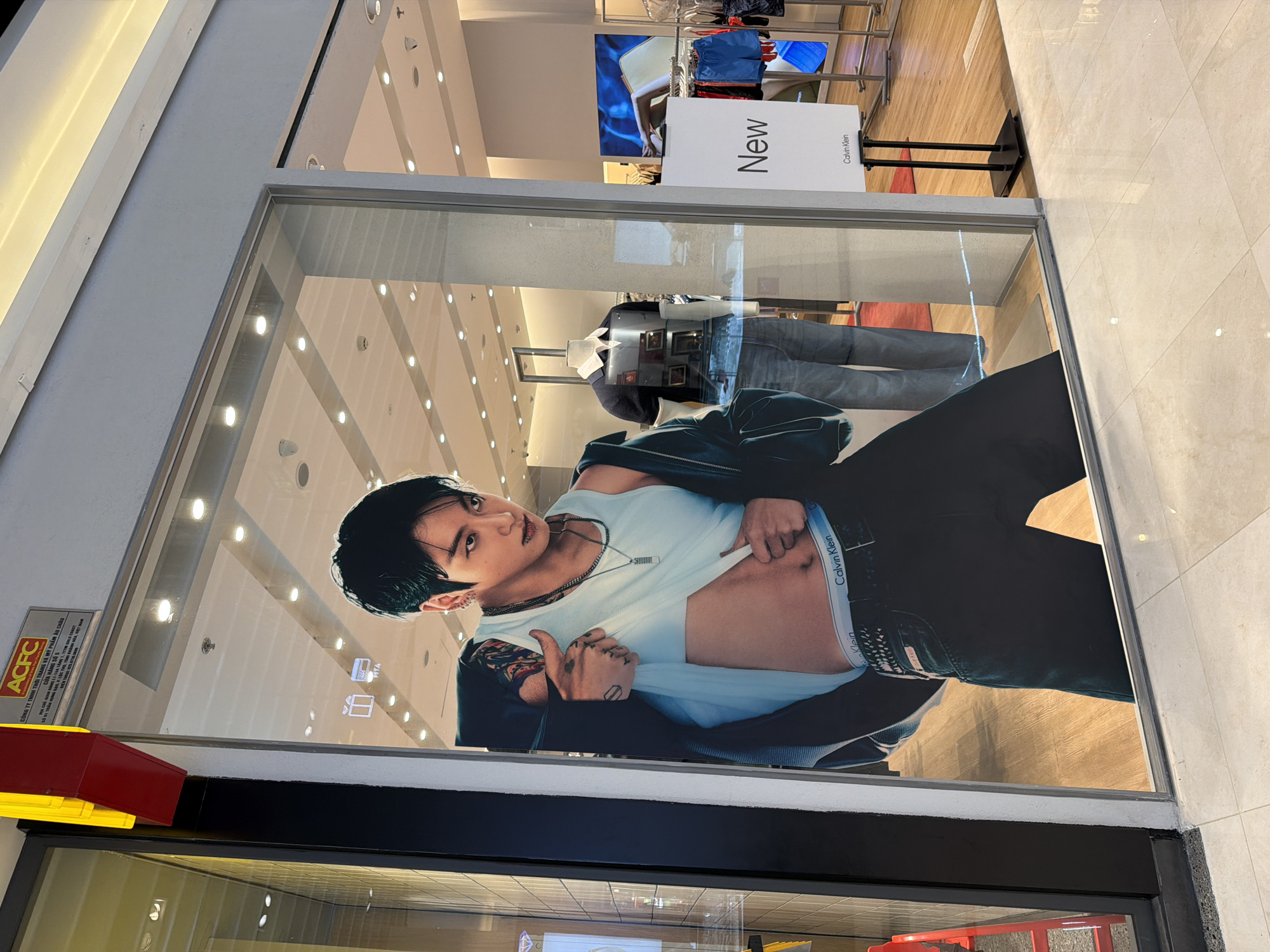The image shows a shop window display in Nha Trang, Vietnam. The display features a large advertisement of a male model wearing a Calvin Klein outfit. The model is shirtless, revealing his abdomen and Calvin Klein underwear. He is wearing a light blue tank top, a dark jacket, and dark jeans. He has tattoos on his arms. To the right of the advertisement, there is a mannequin wearing a blue polo shirt and jeans, and a display stand with the word "New" and the Calvin Klein logo. Clothing racks with various items are visible in the background, as is a screen displaying an image of a person. A sign at the top left of the image reads "ACFC THUONG TRANG MY PHAM AU CHAU". The floor of the shop appears to be made of light-colored tiles.