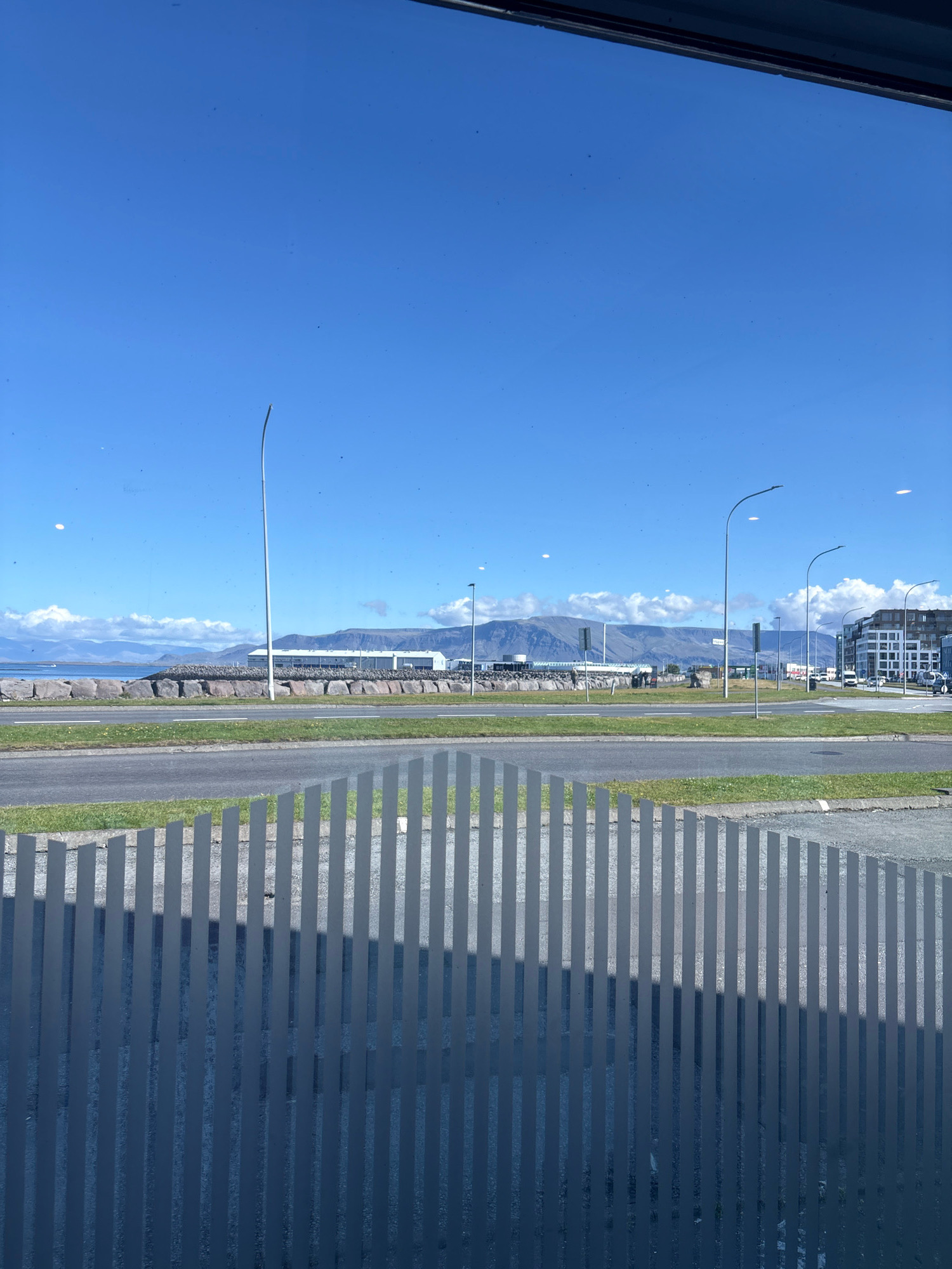 The image shows a view of a street and coastline in Reykjavik, Iceland, from behind a frosted glass partition with vertical stripes. In the foreground, the frosted glass creates a pattern of vertical lines, obscuring a clear view of the area immediately beyond it. The middle ground features a two-lane road with a grassy median strip. Beyond the road, a rocky coastline is visible, with a body of water to the left. In the distance, a long, low mountain range forms the horizon, under a clear blue sky dotted with a few fluffy white clouds. Several modern buildings, including a long, white industrial-style structure and taller office buildings, are situated along the coastline and further inland. Streetlights are visible along the road and near the buildings. There are no people clearly discernible in the image. The overall scene depicts a bright, sunny day in an urban coastal environment.