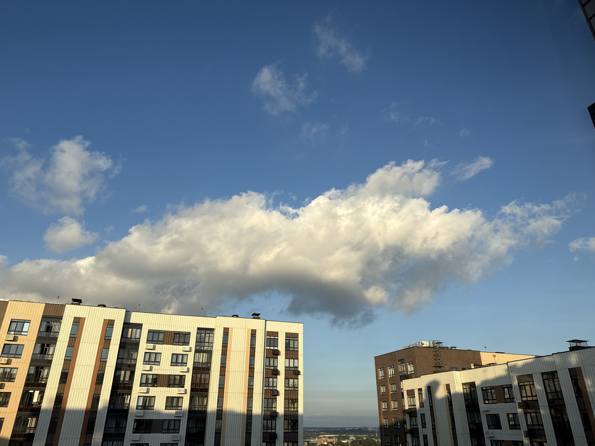 The image depicts a view of modern apartment buildings against a backdrop of a bright blue sky with scattered white clouds. The buildings, featuring contemporary architecture with a mix of light-colored tiled facades and darker accents, are illuminated by the sun, casting shadows across their surfaces. The sky is a prominent element, with a large, voluminous cloud formation dominating the upper central portion of the frame, catching the sunlight and appearing bright white. The scene suggests an urban environment, likely Lyubertsy, Russia, given the context. The image is a still photograph capturing an outdoor scene during daylight hours, likely late afternoon or early evening due to the angle of the sunlight and resulting shadows. There are no people visible in the image. The overall impression is one of a modern cityscape under a dynamic sky.