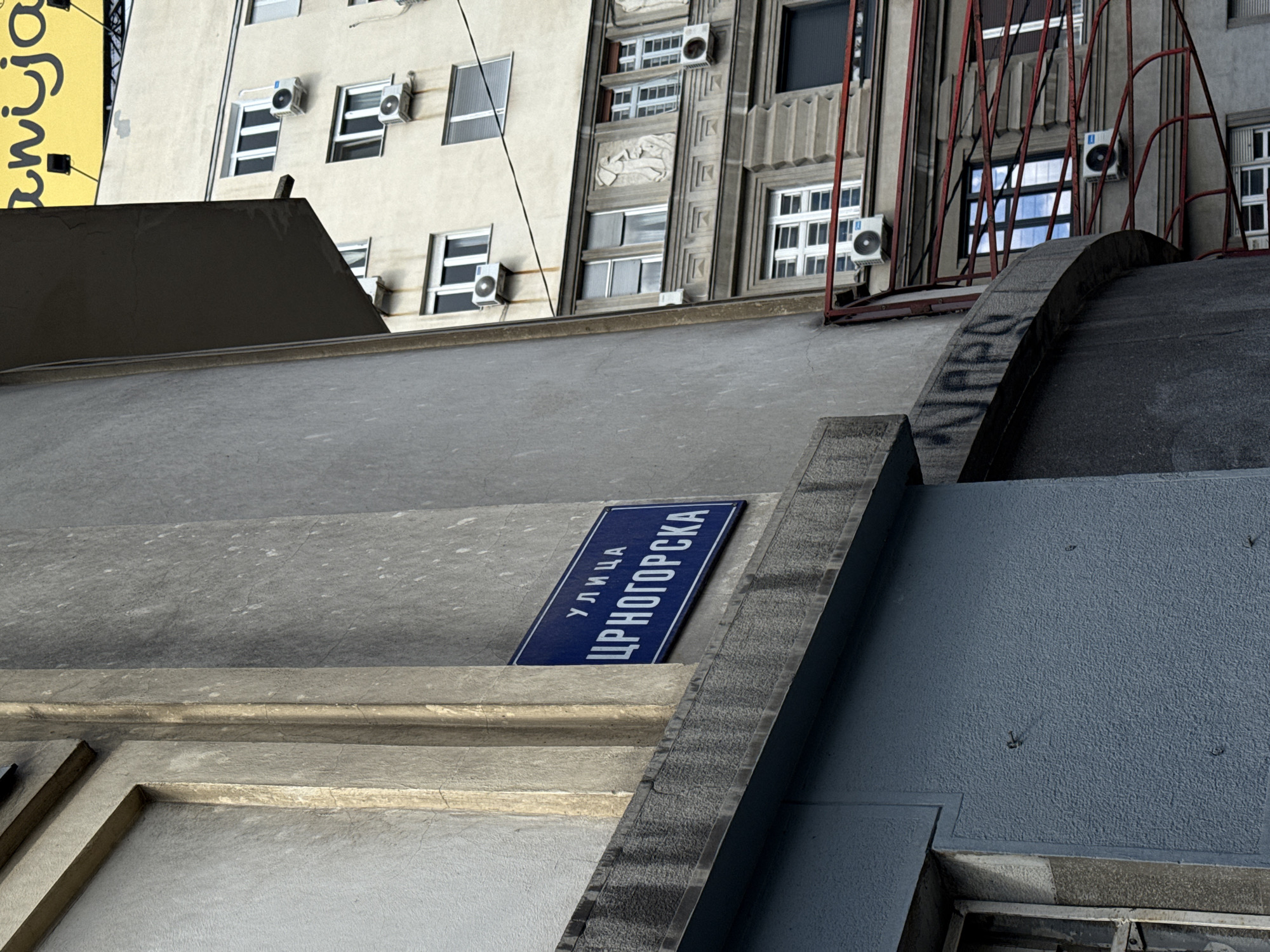 This image captures an upward-angled view of an urban streetscape, likely in Belgrade, Serbia, given the visible Cyrillic text and location hint. The foreground is occupied by various textured concrete or stone architectural elements, suggesting steps, walls, or a building's foundation, with varying shades of grey.
A prominent blue street sign with white Cyrillic text, reading "УЛИЦА ЦРНОГОРСКА" (ULICA CRNOGORSKA), meaning "Montenegrin Street," is positioned diagonally on one of these concrete surfaces.
In the background, two multi-story buildings are visible. The one on the left is light-colored, featuring multiple windows and several white external air conditioning units. The building to its right is darker and more architecturally ornate, with decorative bas-relief sculptures, fluted elements, and a red, somewhat industrial-looking metal structure or railing extending upwards. This building also displays windows and air conditioning units. A partial yellow banner with Cyrillic text is visible on the far left.
The scene appears to be under natural daylight, possibly overcast or late afternoon, as the light is soft and there are no harsh shadows. There are no people visible, and no specific activities are taking place, presenting a static snapshot of an urban environment. The composition emphasizes architectural details, textures, and the unique perspective of looking up from what appears to be a lower vantage point within the city.