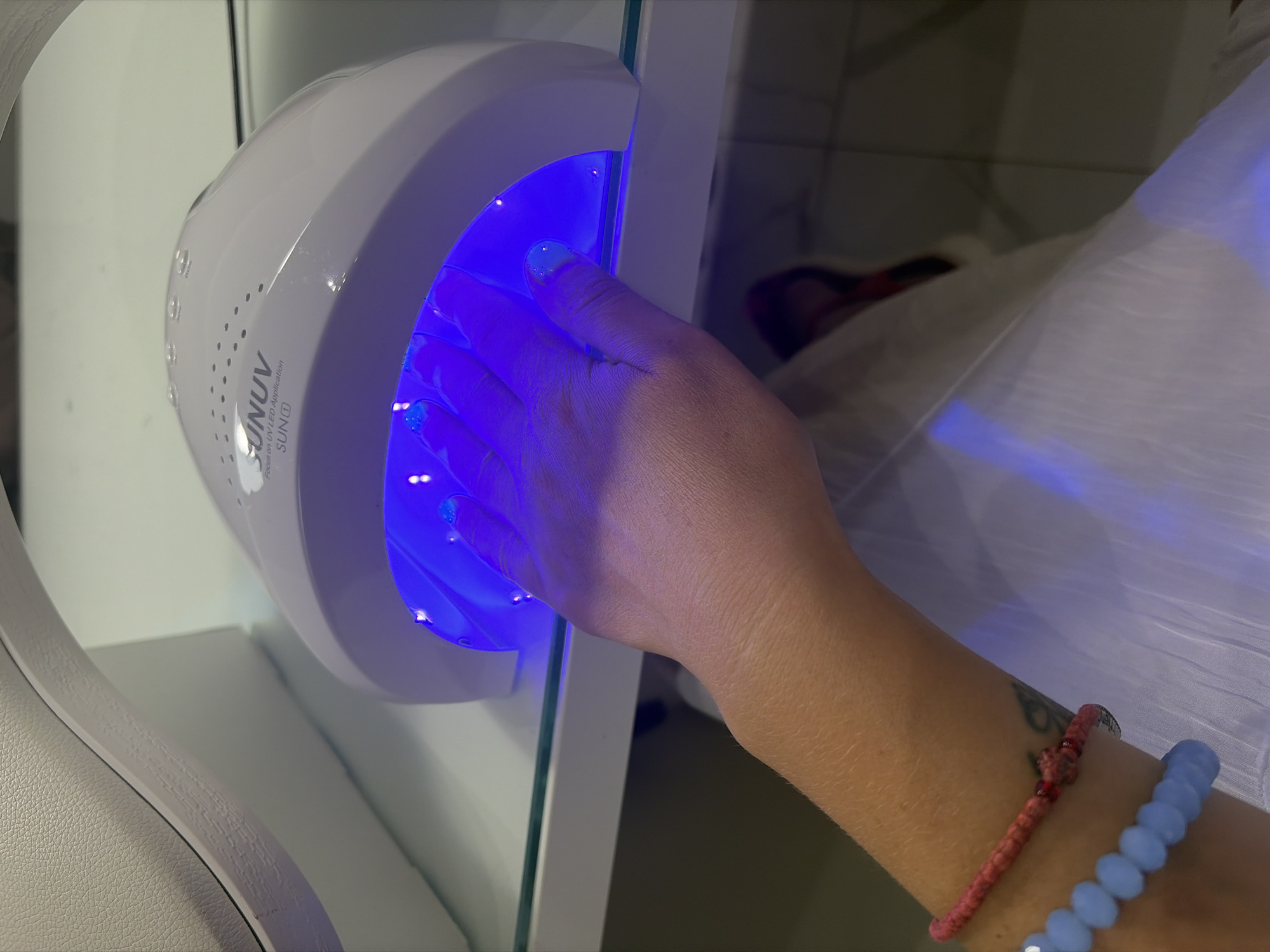 A person's left hand is positioned inside a white SUNUV brand UV/LED nail lamp, which emits a bright blue light. The nails appear to have a light blue gel polish. The hand is adorned with two bracelets: one pink woven bracelet with red beads and a silver charm, and another bracelet composed of light blue opaque beads. The lamp sits on a white surface, possibly a shelf, with a textured light-colored object, possibly a shoe or bag, in the upper left corner. The background is blurred and appears to be an indoor environment. The scene is consistent with a nail salon or a home manicure setup. There is no information in the image to suggest a location in Budva, Montenegro.