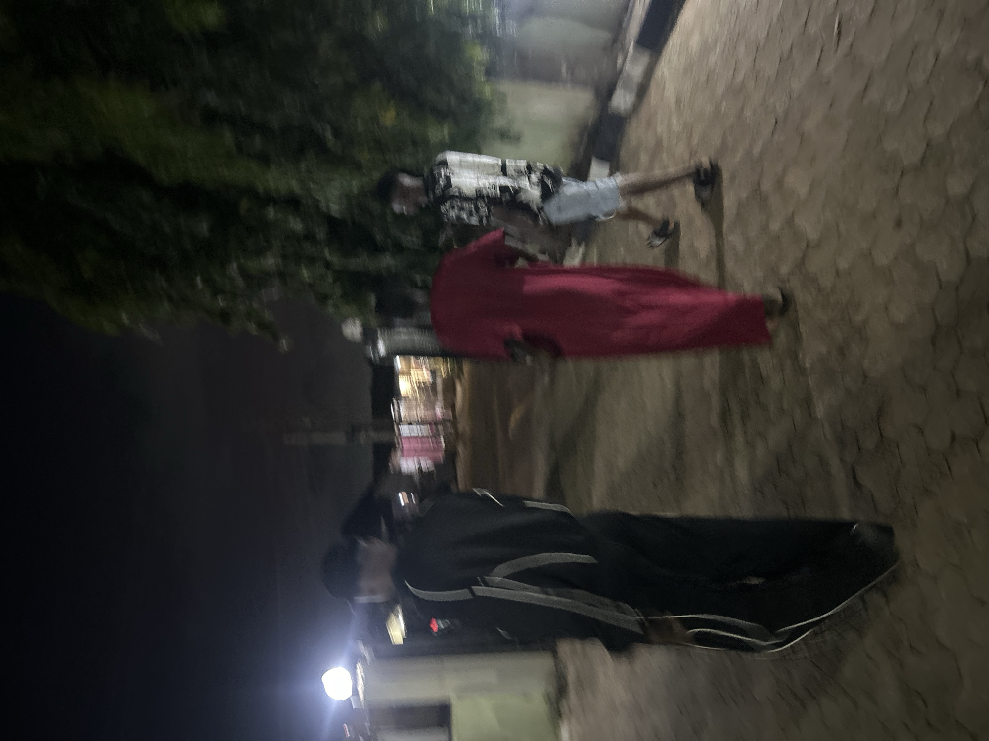The image shows three individuals on a paved surface resembling a sidewalk. One person is wearing a dark jacket and black pants. Another is draped in a red garment, possibly a sheet or cloth. The third person is wearing a light-colored patterned shirt and light-colored shorts. The environment appears to be at night, with streetlights visible in the background, near what looks like a building. There is also foliage on the left side of the image. The location appears to be urban.