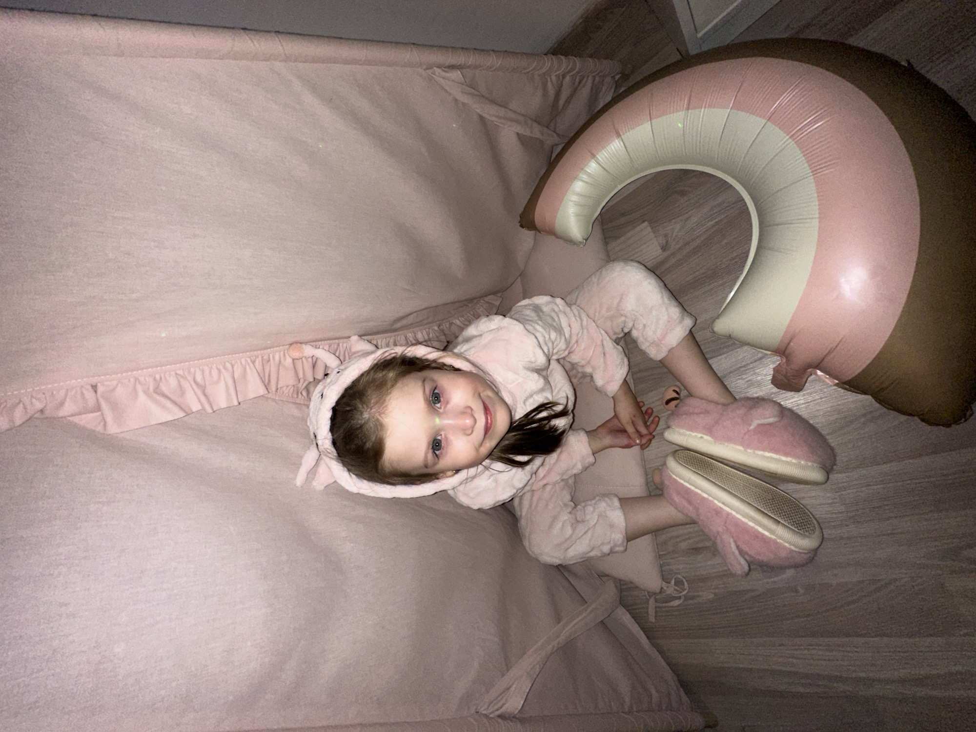 This image, taken in Chaikovsky, Russia, features a young child, likely a girl, sitting indoors. She is positioned centrally, looking upwards towards the camera with a gentle smile, conveying a sense of contentment. The child has fair skin, blue eyes, and light brown hair, which is partially covered by the hood of her attire.
She is dressed in a cozy, pink, fluffy onesie or pajamas, complete with a hooded top that appears to have small ears. Her feet are adorned with matching pink, fluffy slippers, one of which is slightly askew on the wooden floor. The child is seated on a soft, light pink fabric, which forms a cozy, enclosed space, possibly part of a play tent, bed canopy, or a large floor mat. The fabric features ruffled detailing along its upper edge.
Adjacent to the child, on the light-colored wooden floor, lies a large, inflatable object shaped like a rainbow. This toy is striped with shades of brown, pink, and off-white or cream. The overall lighting in the scene is dim, suggesting an evening setting or an indoor space with low light. No text is visible within the image.