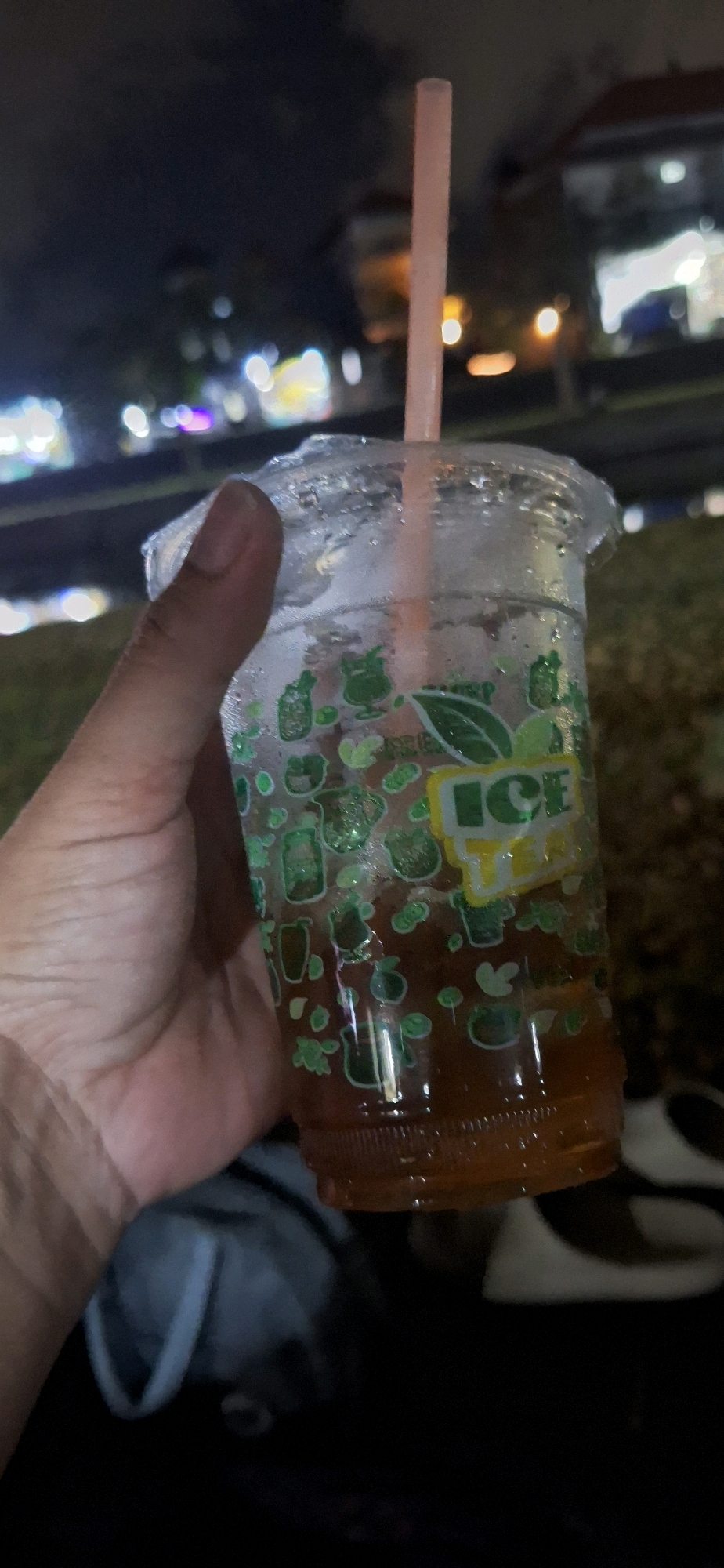 A hand holds a clear plastic cup filled with iced tea, with a pink straw protruding from the top. The cup has a green and yellow logo that reads "ICE TEA" and is decorated with small green drawings of fruit and leaves. The cup is condensation-covered, indicating it contains a cold beverage. The background is blurred and dark, suggesting it is nighttime. In the distance, there are out-of-focus lights, possibly from buildings or streetlights, indicating an urban environment. The ground appears to be covered in gravel or small stones. A portion of a white and black sneaker is visible at the bottom right of the image. The overall scene is consistent with a person holding a drink outdoors at night, possibly in a park or a street area within the city of Denpasar, Indonesia.