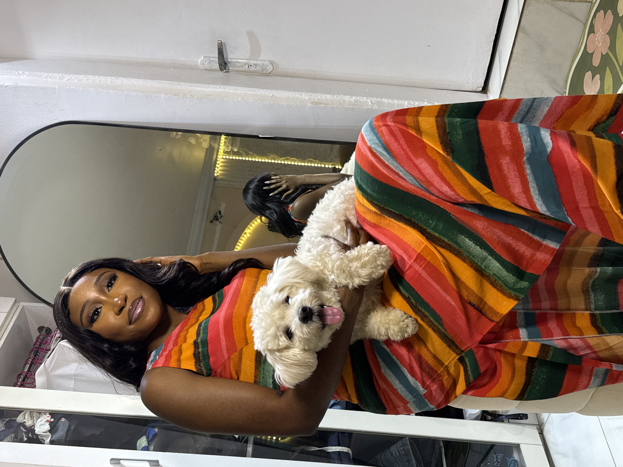 In a room that appears to be within a residential setting in Abuja, Nigeria, a young Black woman with long, dark, wavy hair is seated, holding a small white dog. The woman is wearing a vibrant, sleeveless maxi dress adorned with horizontal stripes in various colors, including red, orange, green, and blue. Her makeup is noticeable, with winged eyeliner and a glossy lip color. The dog, held in her arms, has its tongue out and appears to be panting.
Behind the woman, a large, arch-topped mirror reflects the room. In the mirror's reflection, the woman is partially visible, with her face obscured by her hand, and a string of yellow lights is visible in the background, suggesting ambient lighting or decoration. To the right of the woman, a white door with a silver door handle is present. To the left, a portion of a closet or wardrobe with white doors is visible, containing neatly hung clothes. The floor appears to be tiled. The overall ambiance of the image suggests an indoor, casual setting.