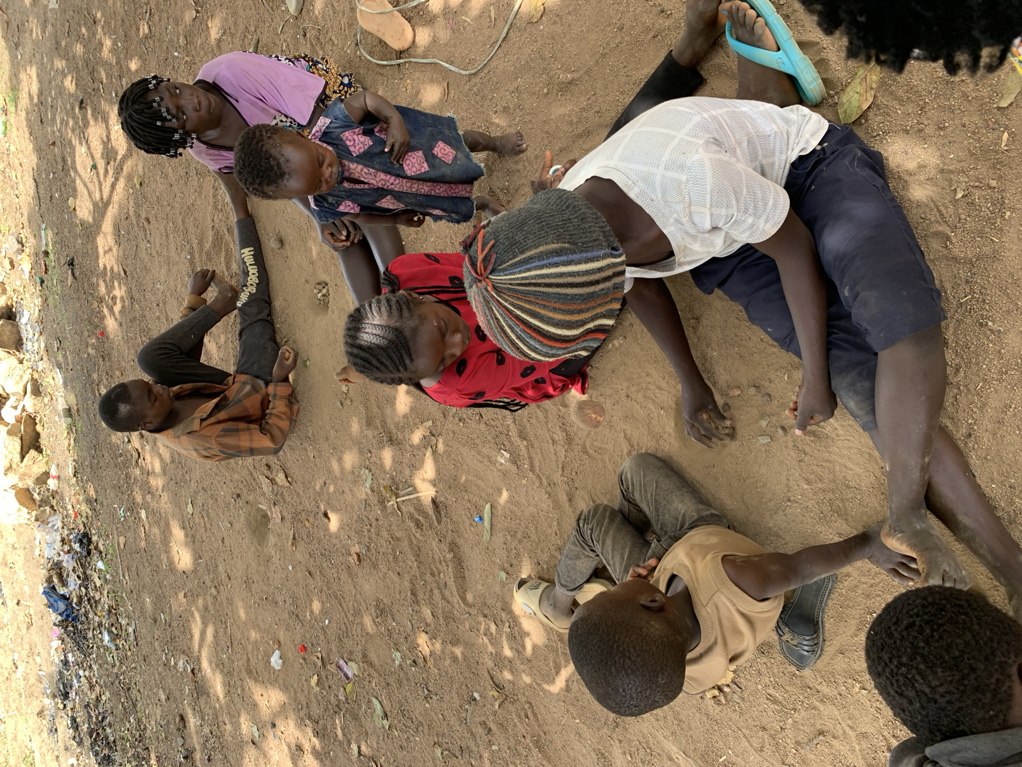 In Rafin Bauna, Nigeria, a group of children are sitting and lying on a dirt ground. At the left, a boy in a plaid shirt has his legs raised. Adjacent to him, a girl wearing a purple shirt and a girl in a patterned dress are seated. A girl with a multicolored hat and red shirt sits next to them. A boy in a white shirt and blue pants is reclining. Another boy in a tan shirt and grey pants is lying down. Debris is scattered in the background.