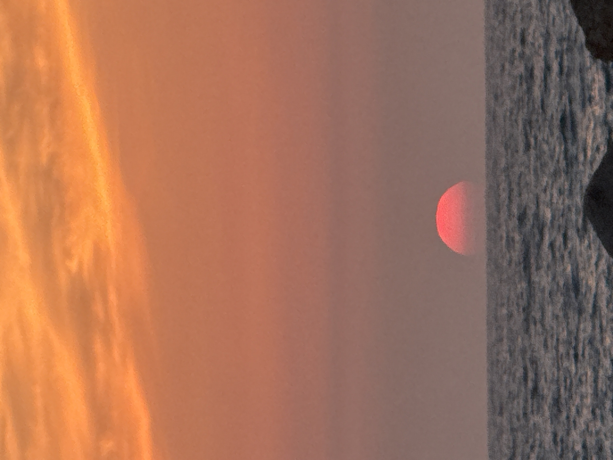 A vertical image captures a sunset over the Mediterranean Sea near Tel Aviv, Israel. To the left is an orange sky with cloud formations. The sky fades to a pale orange and grey closer to the center of the frame, where the red setting sun is visible just above the horizon. To the right of the sun is dark grey-blue sea with turbulent waves crashing against a shoreline, with a darker boulder protruding along the shore.