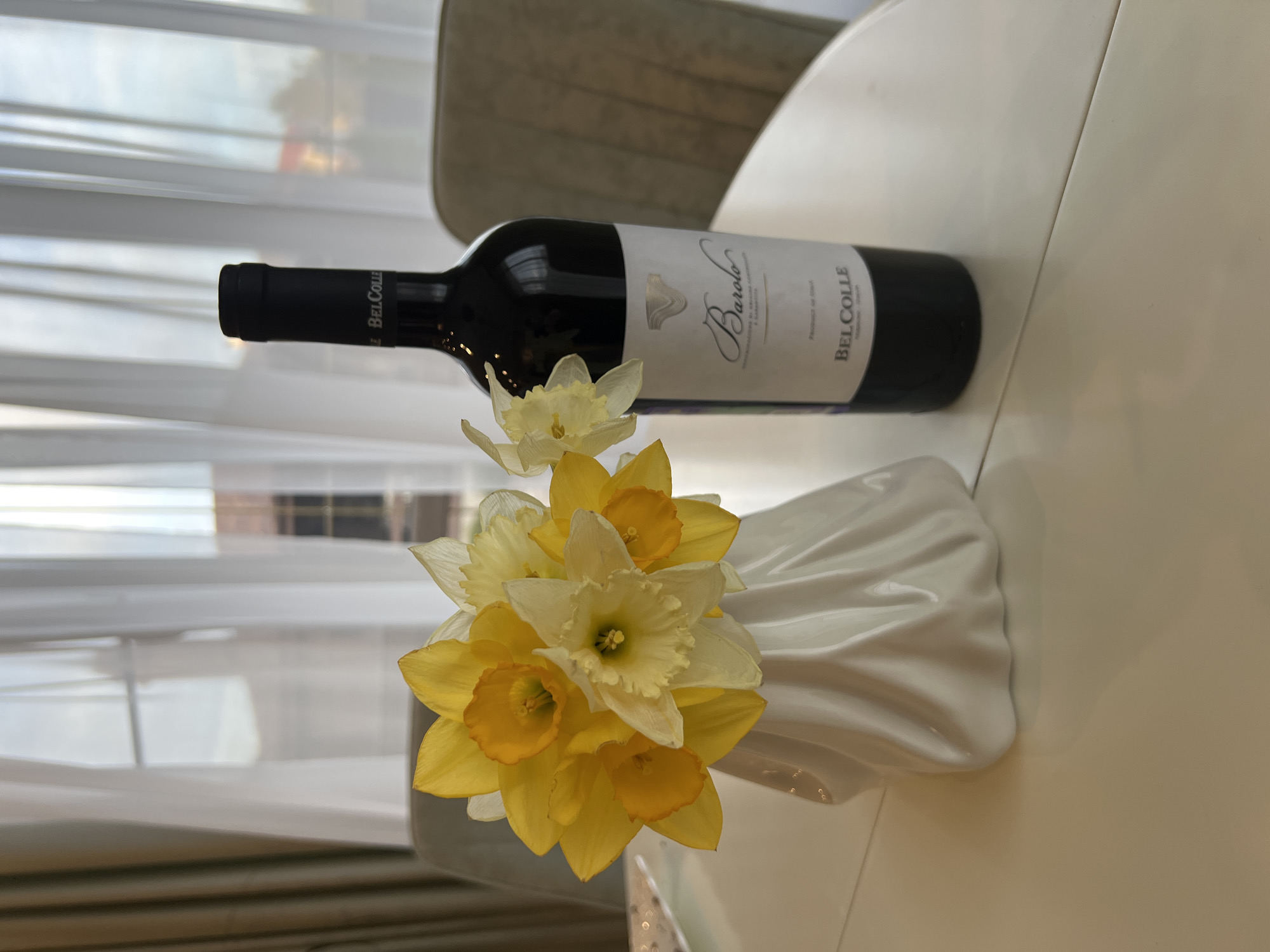 A bottle of Bel Colle Barolo wine lies on a white table, alongside a white vase filled with yellow and white daffodils. The scene is located indoors near a window covered with a sheer white curtain. Parts of a chair are visible.