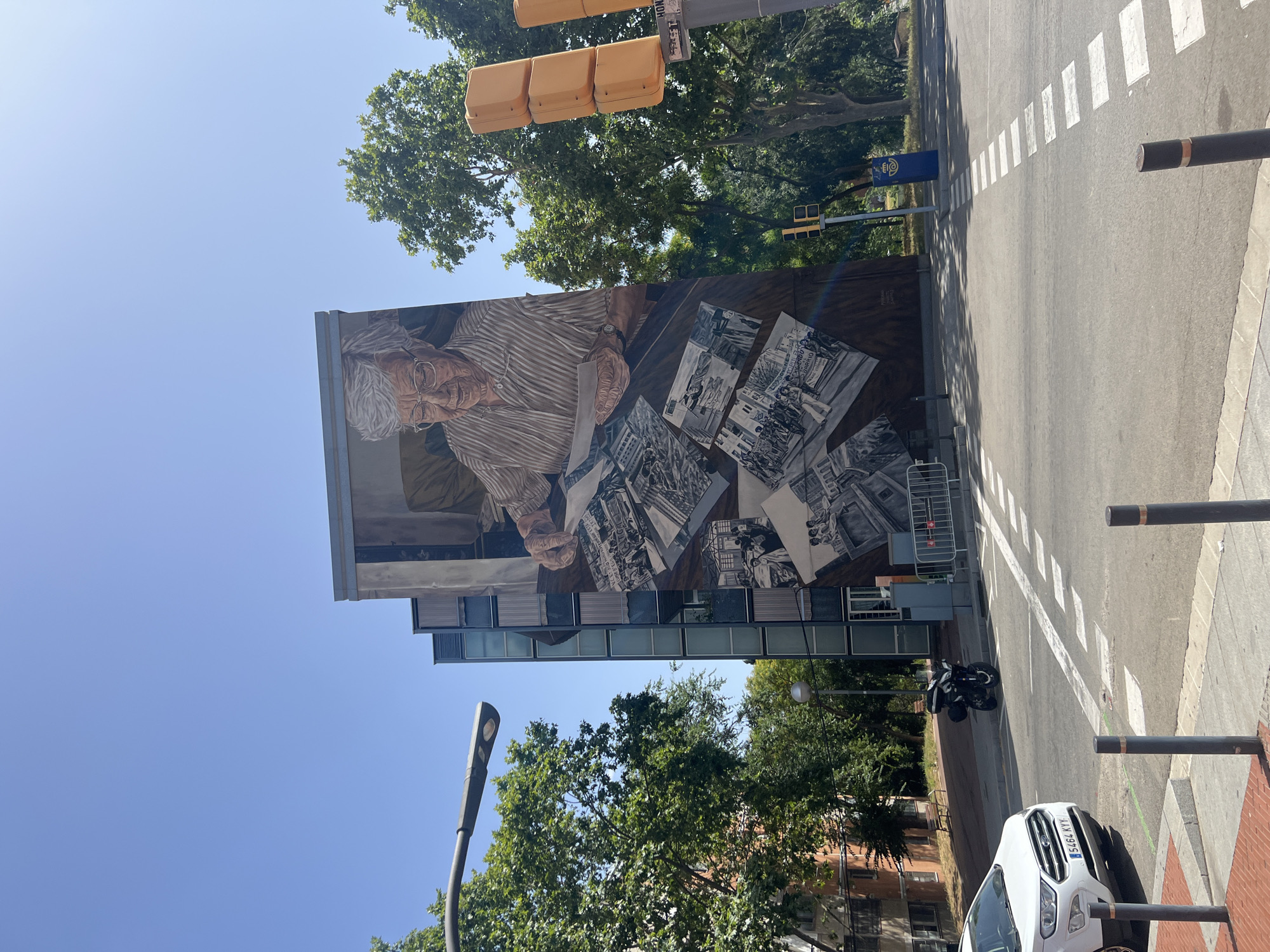 The image shows a large mural on the side of a building in Barcelona, Spain. The mural depicts an elderly woman with gray hair and glasses looking at photographs spread out on a table. The photographs show various scenes, including a crowd of people, buildings, and what appears to be a protest. The building itself is a modern structure with glass windows. In the foreground, there is a street with a white car, a motorcycle parked on the side, and a pedestrian crossing. Traffic lights and street lamps are also visible. The sky is clear and blue, and trees are scattered throughout the scene.