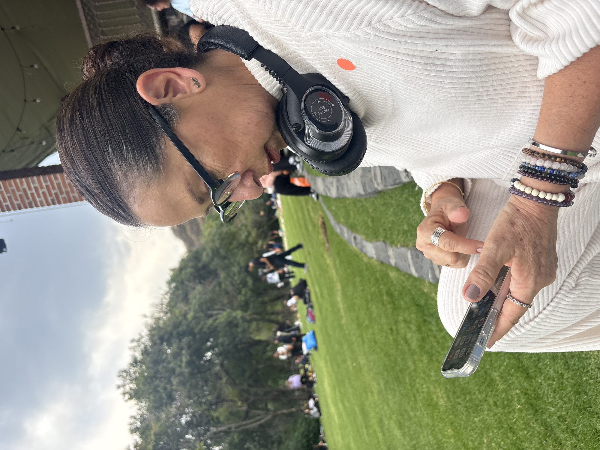 An older adult female is seated outdoors, looking at a smartphone. She wears glasses, headphones, and several bracelets on her right wrist. The woman wears a white ribbed sweater. In the background, several people are seated on a grassy lawn under trees, possibly in Santa Ana Acozautla, Mexico. A walkway is visible to the right.