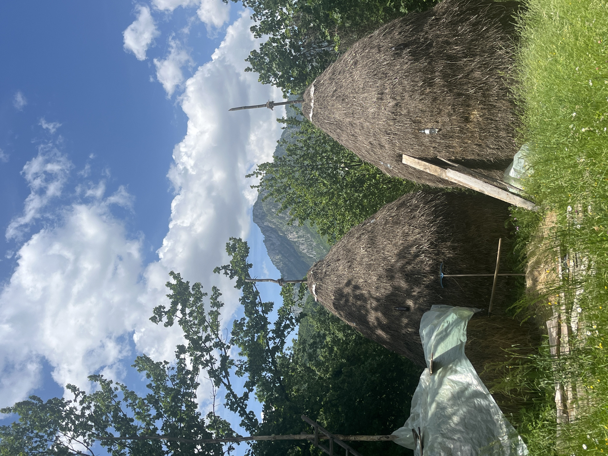 The image depicts a rural scene in Kaludra, Montenegro. Two conical haystacks dominate the foreground, constructed of dried grasses or straws. A wooden pole protrudes from the top of each stack. One haystack is partially covered with a translucent plastic sheet. In the background, a mountain peak is visible amidst lush green trees and vegetation. The sky is blue with scattered white clouds. A pickaxe and other tools are placed on the ground near the haystacks. The image is rotated ninety degrees clockwise.
