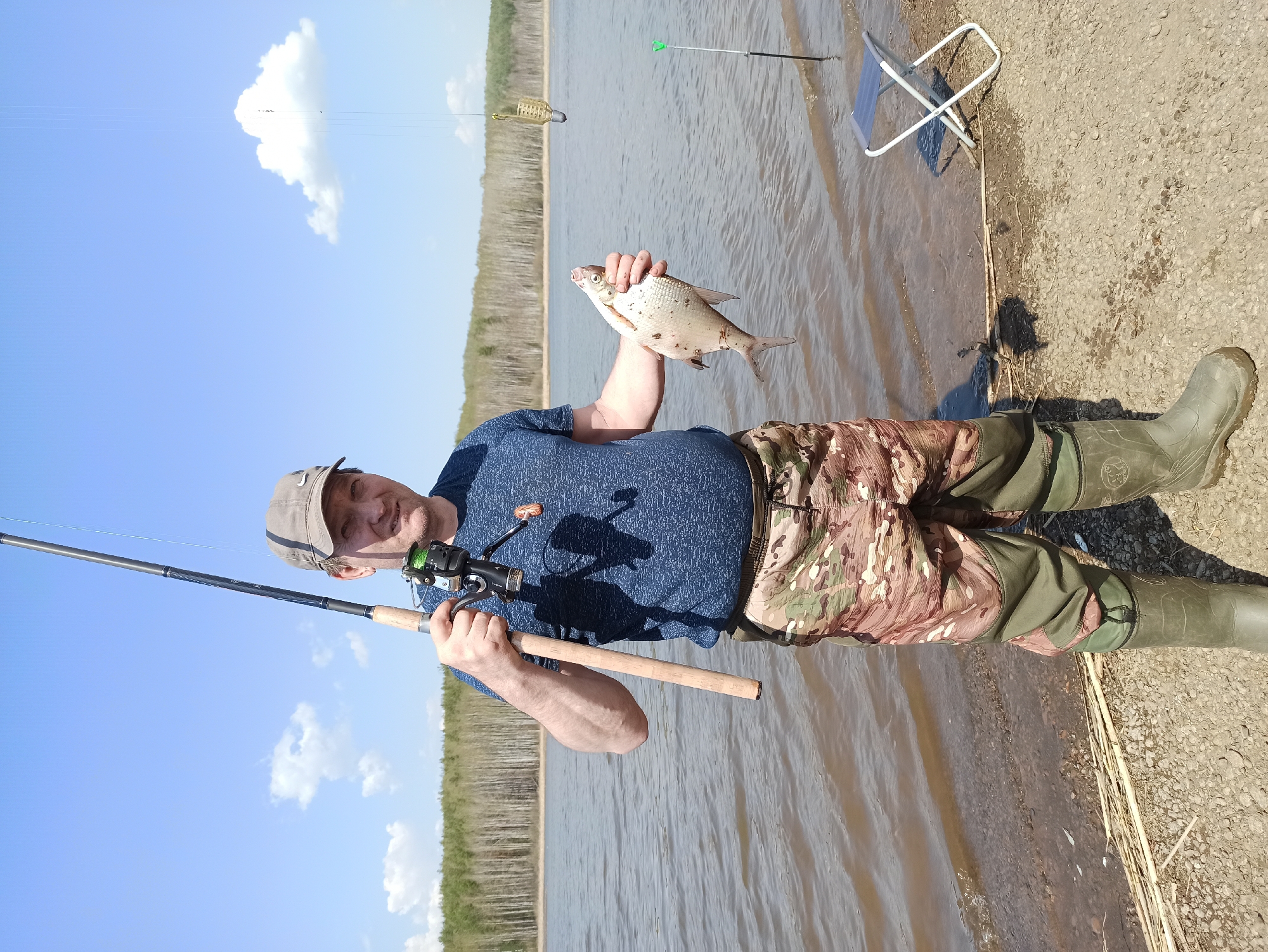 A man stands at the edge of a body of water, holding a fish in his right hand and a fishing rod in his left. He wears a blue shirt, camouflage pants, a cap, and green rubber boots. Behind him, there is a folding chair. In the water, there are fishing lines and equipment. The background shows trees and a partly cloudy sky. The location is the city of Unknown, Unknown.