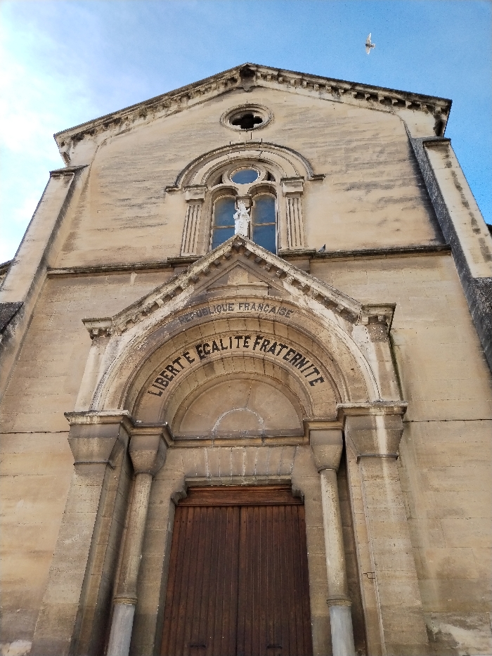 A low-angle shot reveals a stone building under a partly cloudy sky. Above the wooden double doors is an arched stone inscription "LIBERTE EGALITE FRATERNITE" beneath "REPUBLIQUE FRANCAISE". Stone columns frame the entrance. A niche with a statue sits above the entrance, topped by a circular window. The building's facade is textured, and a single bird flies in the sky.