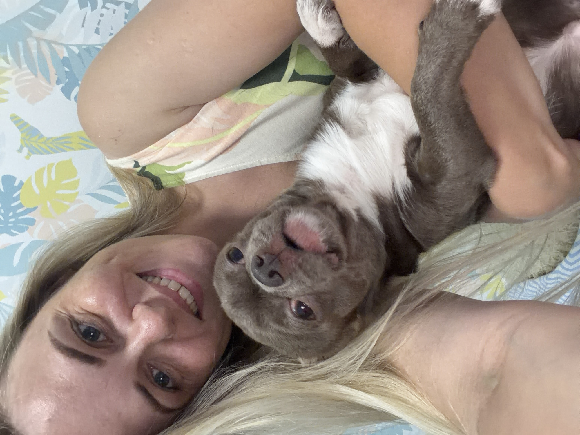 In this close-up, upside-down selfie, a blonde woman lies on her side, smiling at the camera. She is embracing a light gray and white dog, which is also upside down and appears to be looking at her. The woman is wearing a light-colored tank top with a tropical leaf pattern in green, peach, and yellow. Her hair is long and blonde, fanned out around her. The dog has a short coat, with distinct gray patches on its body and head, and a white chest and muzzle. Its ears are floppy, and its eyes are dark.
The background is a soft, patterned fabric, possibly a bedspread or blanket, with a light blue and white striped design interspersed with yellow and blue floral or leaf motifs. The overall lighting suggests it's daytime indoors. The scene conveys a feeling of warmth, affection, and intimacy between the woman and her dog. There is no visible text in the image. The location context of Votkinsk, Russia, is not directly discernible from visual cues in the image.