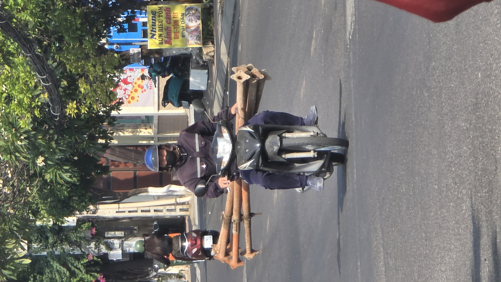 A medium shot of a man riding a motorcycle. In the city of Bắc Nha Trang, Vietnam, a man wearing a blue helmet and a black face mask drives a black motorcycle down a gray street. On the motorcycle are three long, orange-rust-colored metal poles. Another motorcycle follows the man, driven by a person obscured by trees and a wall. Trees are seen on the left side of the frame. In the background are buildings and signs in Vietnamese.