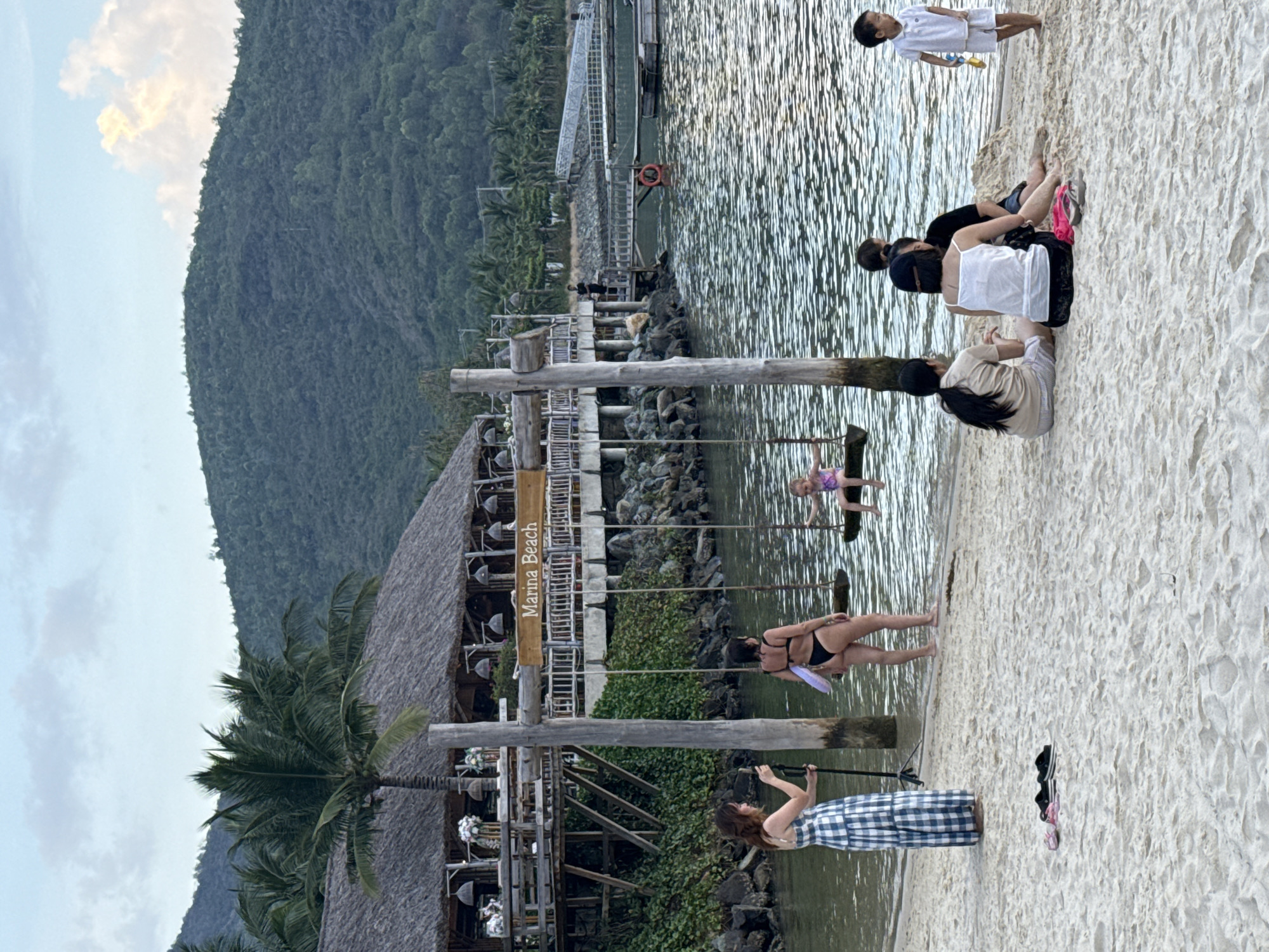 The image depicts a scene at Marina Beach in Bắc Nha Trang Ward, Vietnam. A woman in a blue and white checkered dress is taking a photo of a woman in a black bikini on the beach. A child in a purple swimsuit is on a swing. Other people are sitting on the beach; one child in a white shirt is standing in the distance. There is a restaurant with a thatched roof on the beach. The beach is sandy and there is a mountain in the background.