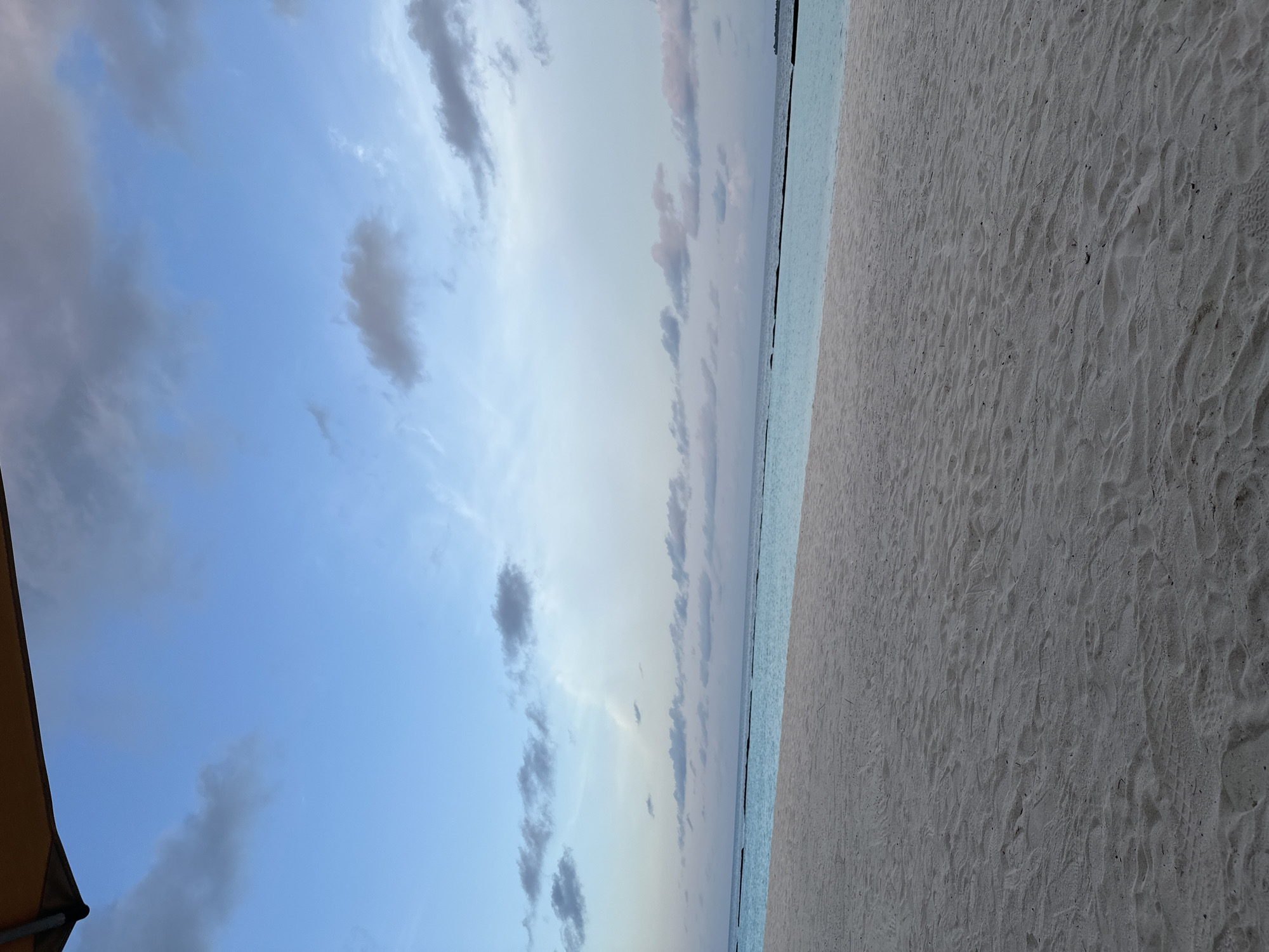 The image shows a tranquil beach scene in Halaveli, Maldives. The foreground is dominated by a wide expanse of light-colored sand, showing gentle undulations and the subtle imprints of footsteps. In the middle ground, a calm, turquoise ocean stretches towards the horizon. A dark, thin line, possibly a barrier or a distant structure, marks the boundary between the water and the horizon. The sky above is a gradient of pale blue, with scattered, wispy clouds in shades of gray and white. The top edge of the frame is partially obscured by the edge of an orange awning or umbrella, casting a slight shadow. No people are visible in the scene. The overall atmosphere is serene and peaceful, indicative of a quiet beach at dusk or dawn.
