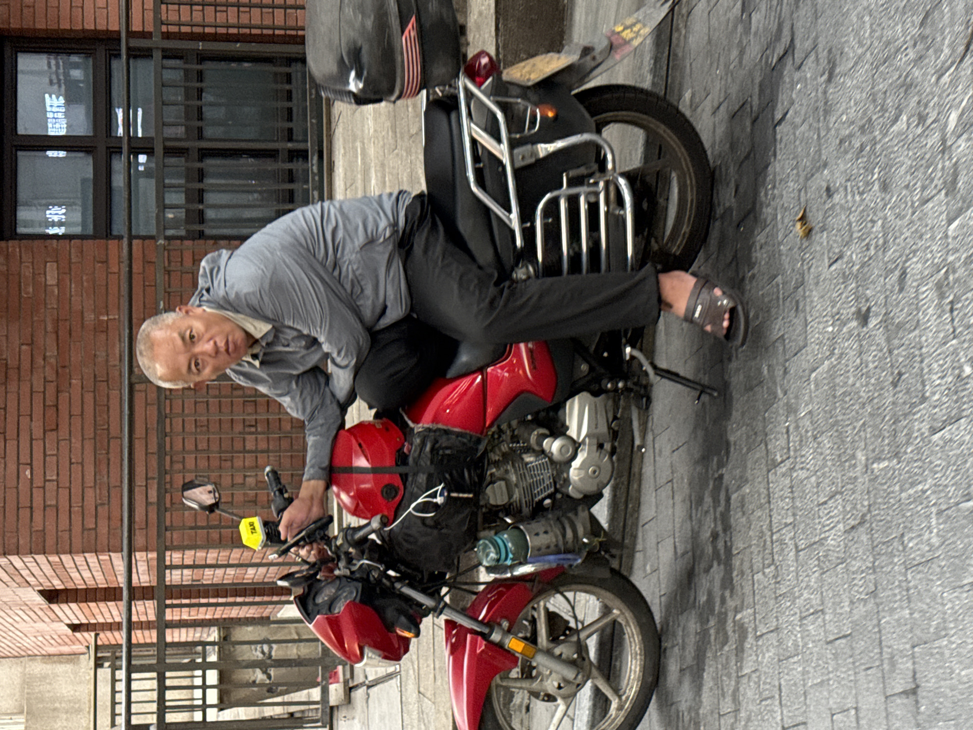 A middle-aged East Asian man with short, graying hair and a light complexion is seated on a red motorcycle. He is wearing a gray hooded jacket over a light-colored shirt and black pants, with dark sandals on his feet. The motorcycle has a black storage box mounted on the rear, a red helmet secured to the back of the seat, and a blue water bottle attached to the side. The man is looking directly at the camera with a neutral expression, his left hand resting on the handlebars and his right hand on his lap. The motorcycle is parked on a brick-paved street, with a metal railing and a brick building in the background. The setting appears to be an urban environment in the Yuzhong District of China.
