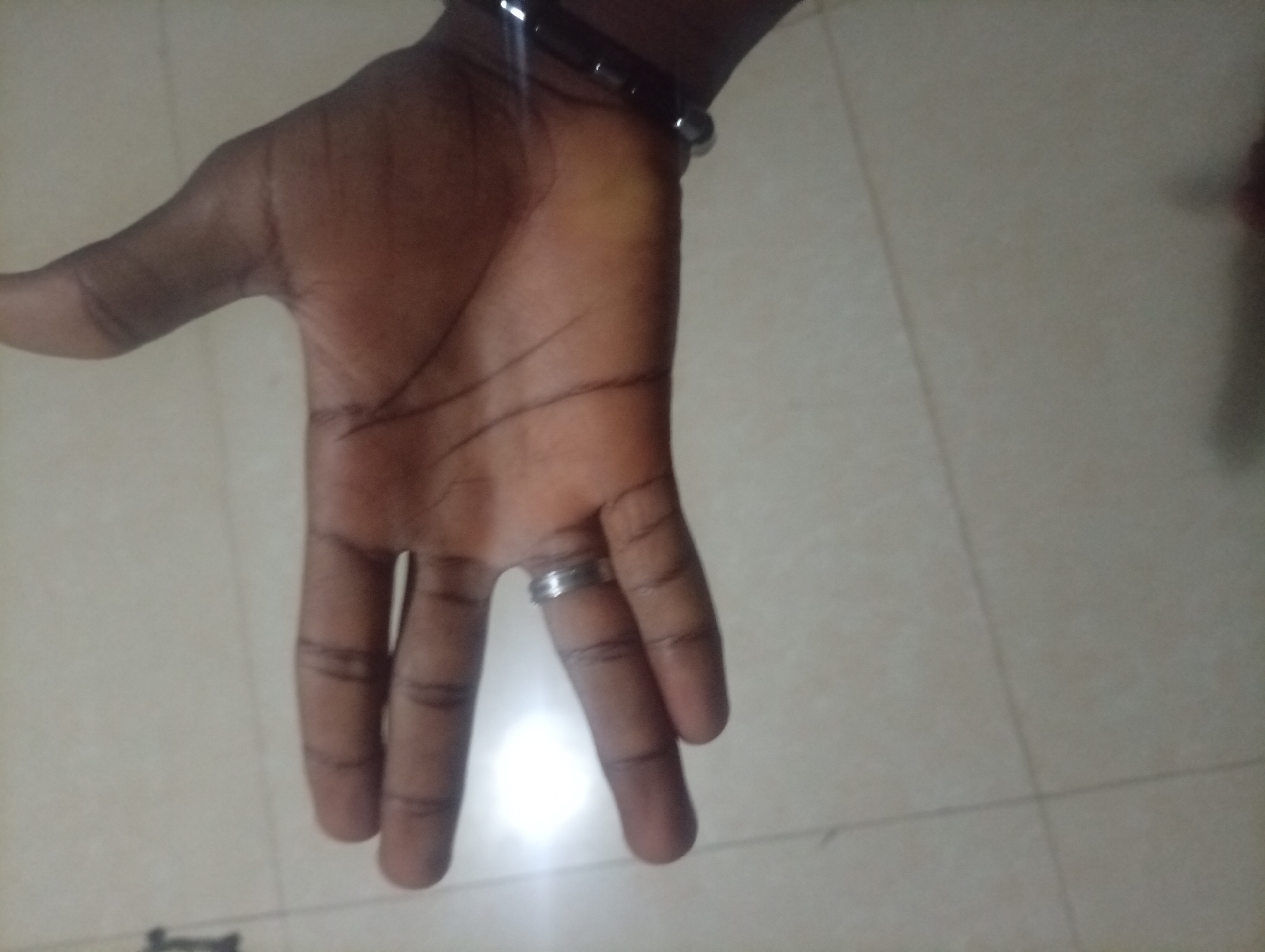 An open left human hand is displayed against a light brown tile floor. The subject wears a silver-colored band on the ring finger. A dark-colored bracelet adorns the wrist. The location is unknown.