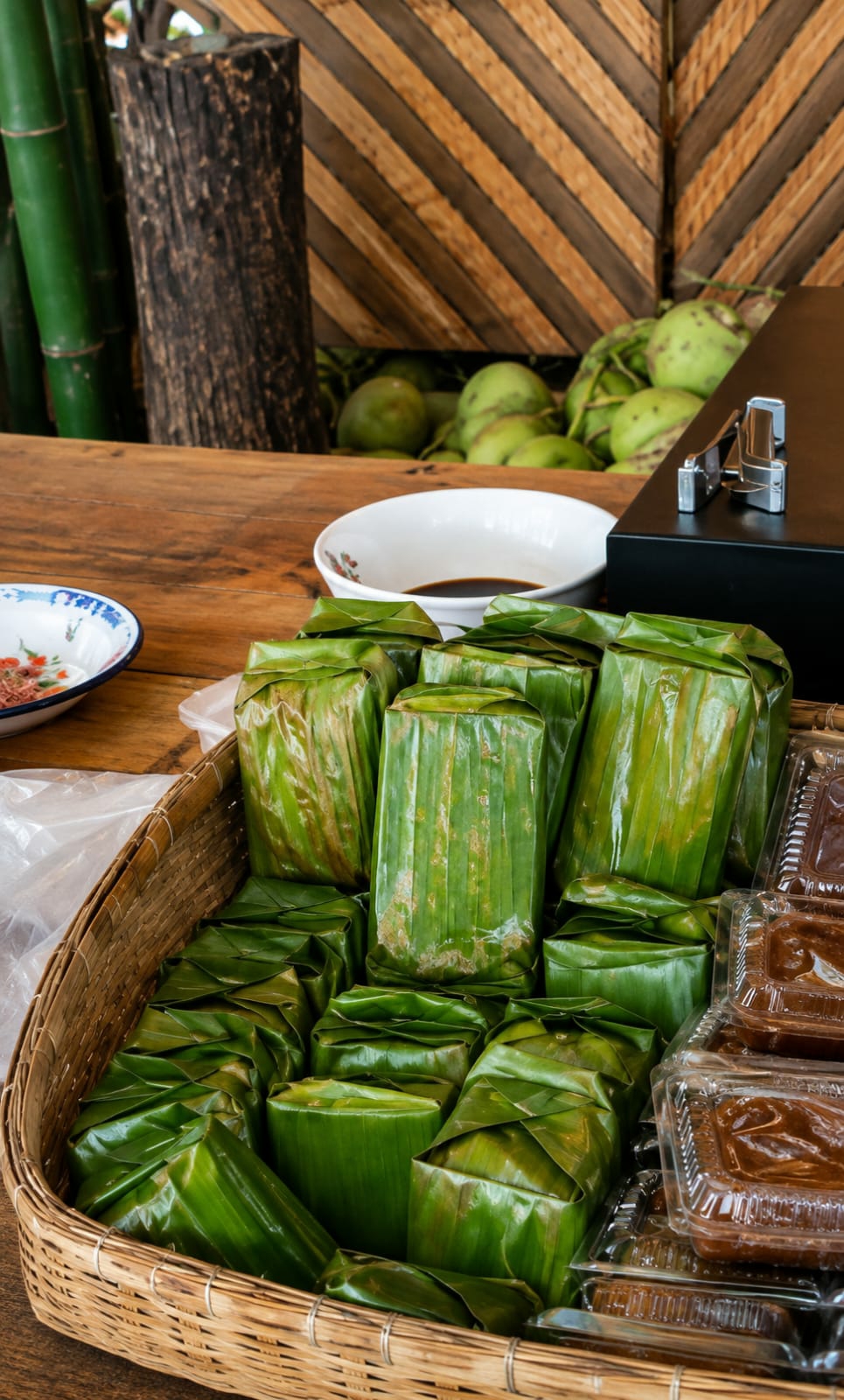 This is a close-up, eye-level shot of a woven basket filled with food items wrapped in banana leaves. The basket is placed on a wooden table. In the foreground, the basket overflows with rectangular parcels wrapped in fresh, green banana leaves, some of which show slight browning. To the right of the basket, several clear plastic containers are stacked, each containing a brown, paste-like food item, possibly a dessert or sauce. In the background, on the same wooden table, there is a white bowl with a dark liquid and a smaller, decorative bowl with a floral pattern. Behind the table, a dark wooden post stands next to several thick, green bamboo stalks. Further back, a rustic wooden wall with diagonal planks forms the backdrop, and a pile of green coconuts is visible behind the wall. The overall impression is of a food stall or market setting, likely in a tropical region. The lighting is natural, suggesting an outdoor or well-lit indoor environment. Based on the visual cues such as the bamboo, coconuts, and the style of food preparation, this scene is characteristic of markets found in the City of Medan, Indonesia, which is known for its tropical climate and diverse culinary traditions.