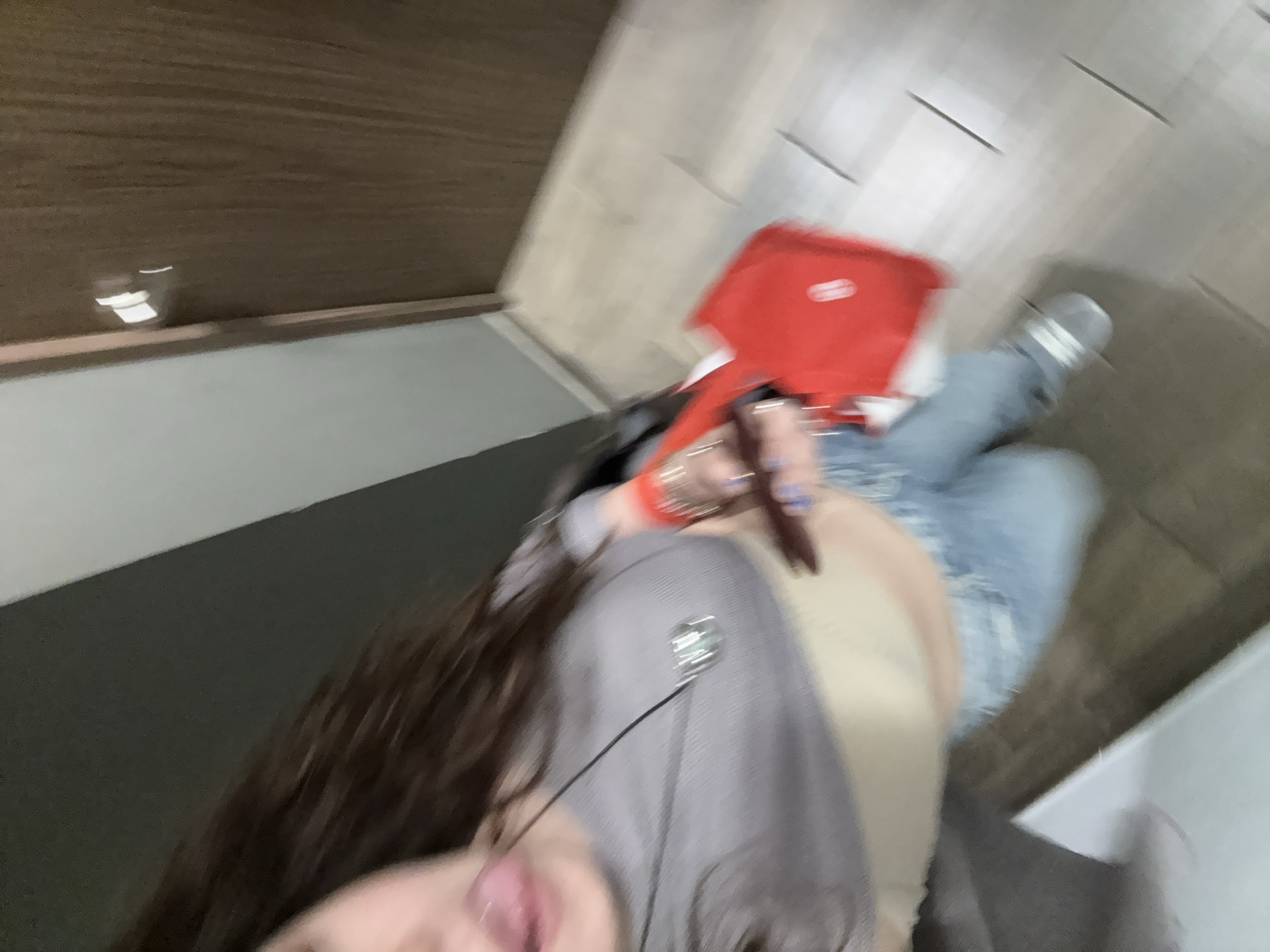 A blurry, close-up, and angled photograph shows a person with long brown hair, wearing a grey top and light-wash denim jeans. They are sticking their tongue out towards the camera. Their right hand is visible, holding a dark object and wearing multiple bracelets on the wrist. A red tray is partially visible in the background. The location appears to be indoors, with a wall and flooring visible in the background. The specific location in Mexico City, Mexico, cannot be determined from the image.