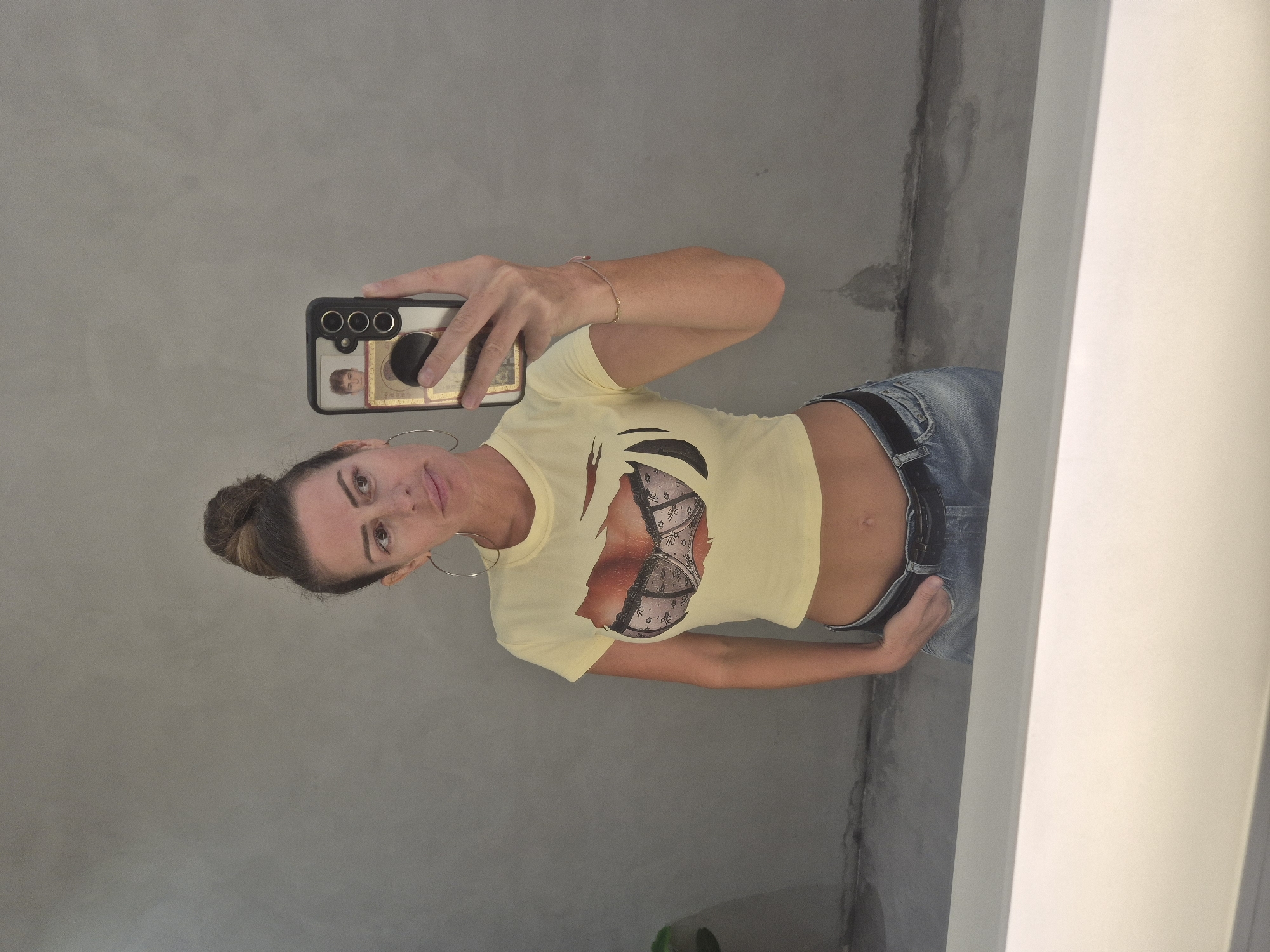 A woman is captured in a mirror selfie, standing upright and looking directly at her reflection (and thus the camera). She appears to be in her 30s or 40s, with her hair styled in a neat bun and wearing large hoop earrings. She has a neutral to slightly smiling expression.
She is wearing a light yellow, short-sleeved crop top that features a prominent graphic of a lacy bra and décolletage. Her midriff is visible above a pair of blue denim jeans, secured with a black belt. A thin bracelet is visible on her right wrist.
The woman is holding a black smartphone with a pop socket in her right hand, positioned to take the mirror selfie. A reflection on her phone screen shows a partial image of a person, possibly a child, framed within what appears to be a picture on the phone's display.
The setting behind her is a plain, light grey wall, which shows some dark vertical streaks and splotches, possibly water stains or signs of wear. To the right of the frame, a stark white vertical architectural element, likely a door frame or partition, indicates an indoor environment. The lighting is bright and even, suggesting a well-lit interior space. The overall scene depicts a casual moment, potentially in a residence or public restroom in Dubai, United Arab Emirates, given the location context.