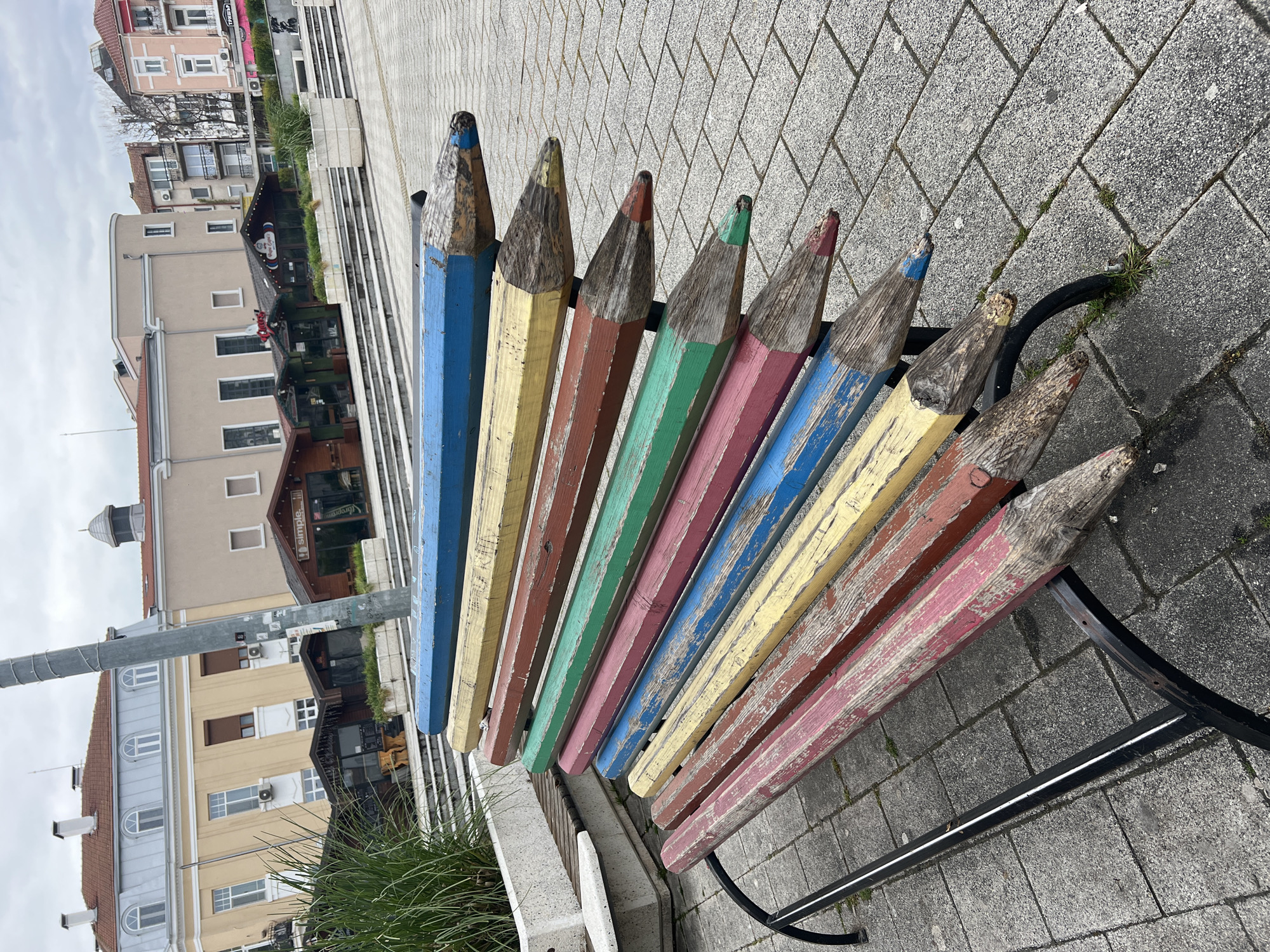 A public bench with a backrest made of large, painted pencils is located in Burgas, Bulgaria. The pencils are arranged side by side, varying in color: blue, yellow, brown, green, pink, and red. The bench's support structure is made of black metal. The bench sits on a paved area composed of gray bricks. In the background, there are buildings and a streetlamp. The sky is overcast.