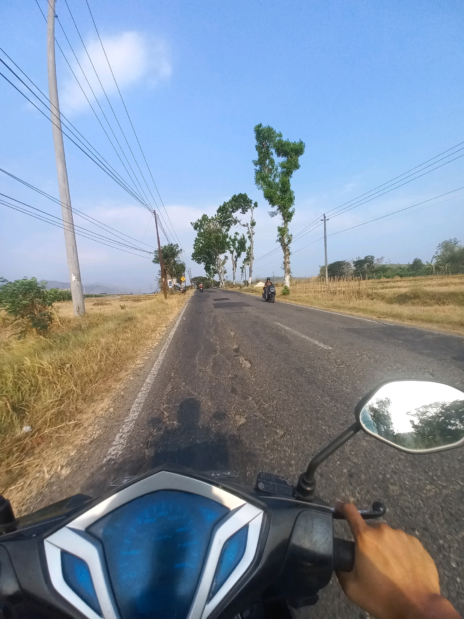 The image depicts a point-of-view shot from a motorcycle traveling on a rural asphalt road in Baturetno, Indonesia. The road has visible cracks and is bordered by dry grass and sparse vegetation on the left, and cultivated fields on the right. Several utility poles with overhead power lines are present along the road. Tall, slender trees with green foliage are spaced along the side of the road. Two motorcyclists are visible on the road ahead, traveling in the same direction as the viewpoint. In the foreground, the handlebars, a portion of the motorcycle's dashboard with blue accents, and a person's right hand on the throttle are visible. The sky is clear blue with a few scattered clouds. The overall scene suggests a daytime journey through a semi-arid rural landscape.