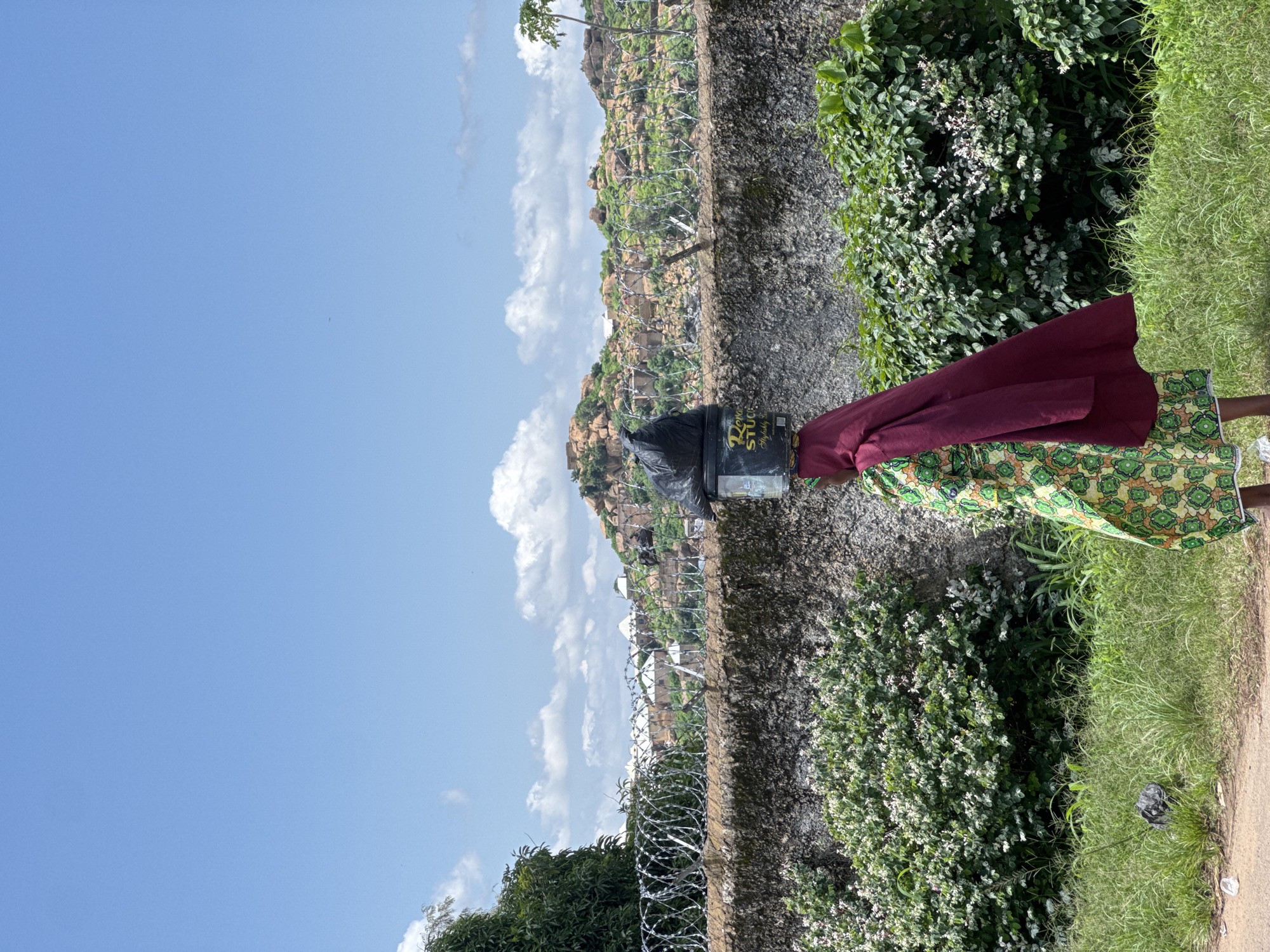 A person walks with a black plastic bag and a cylindrical container balanced on their head along a grassy path beside a stone wall in Jos, Nigeria. The person is wearing a green and yellow patterned dress and a burgundy headscarf. Behind the wall, barbed wire stretches across the landscape, with rocky hills and scattered buildings in the distance under a bright blue sky with white clouds. Lush green bushes with white flowers grow along the base of the wall, and tall grass covers the foreground.