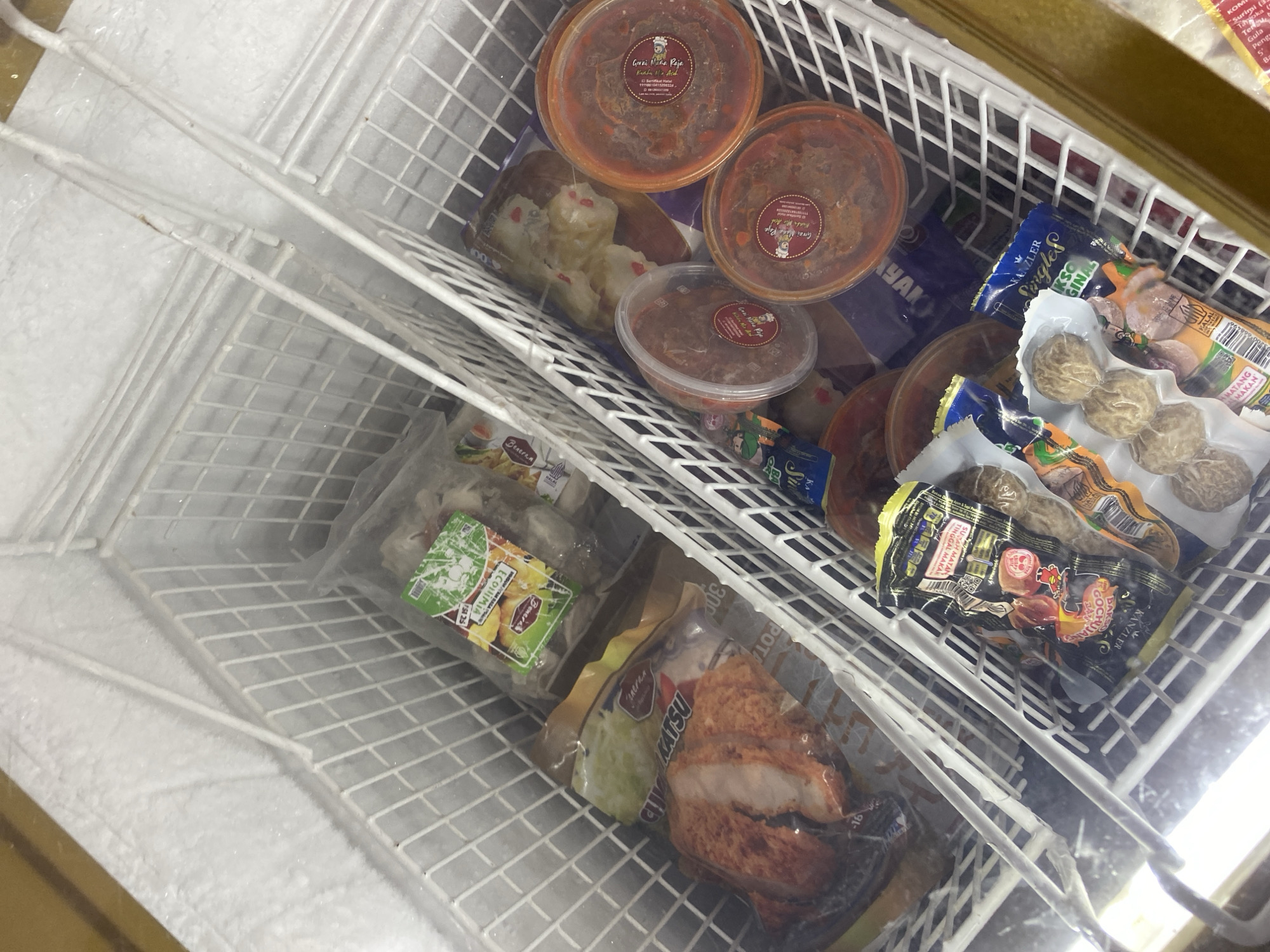 The image shows the interior of a commercial freezer chest filled with various frozen food items. The freezer is divided into two compartments by white wire racks. In the left compartment, there are several plastic-wrapped packages of food. One package, in the foreground, contains what appears to be fried chicken cutlets. Behind it, a package labeled "CHICKEN KATSU" is visible. Further back in this compartment are more plastic-wrapped items.
The right compartment also contains several frozen food products. There are round plastic containers with red lids, three of which are clearly visible and appear to contain a chili paste or sauce. Behind these containers, there are packages of frozen food, including one with small, round meatballs and another with what looks like processed shrimp or dumplings. The freezer interior is coated with frost, indicating it is functioning at a low temperature. The overall scene depicts a commercial storage of frozen food. There are no people or active interactions visible in the image. The location is identified as Payatieng, Indonesia.