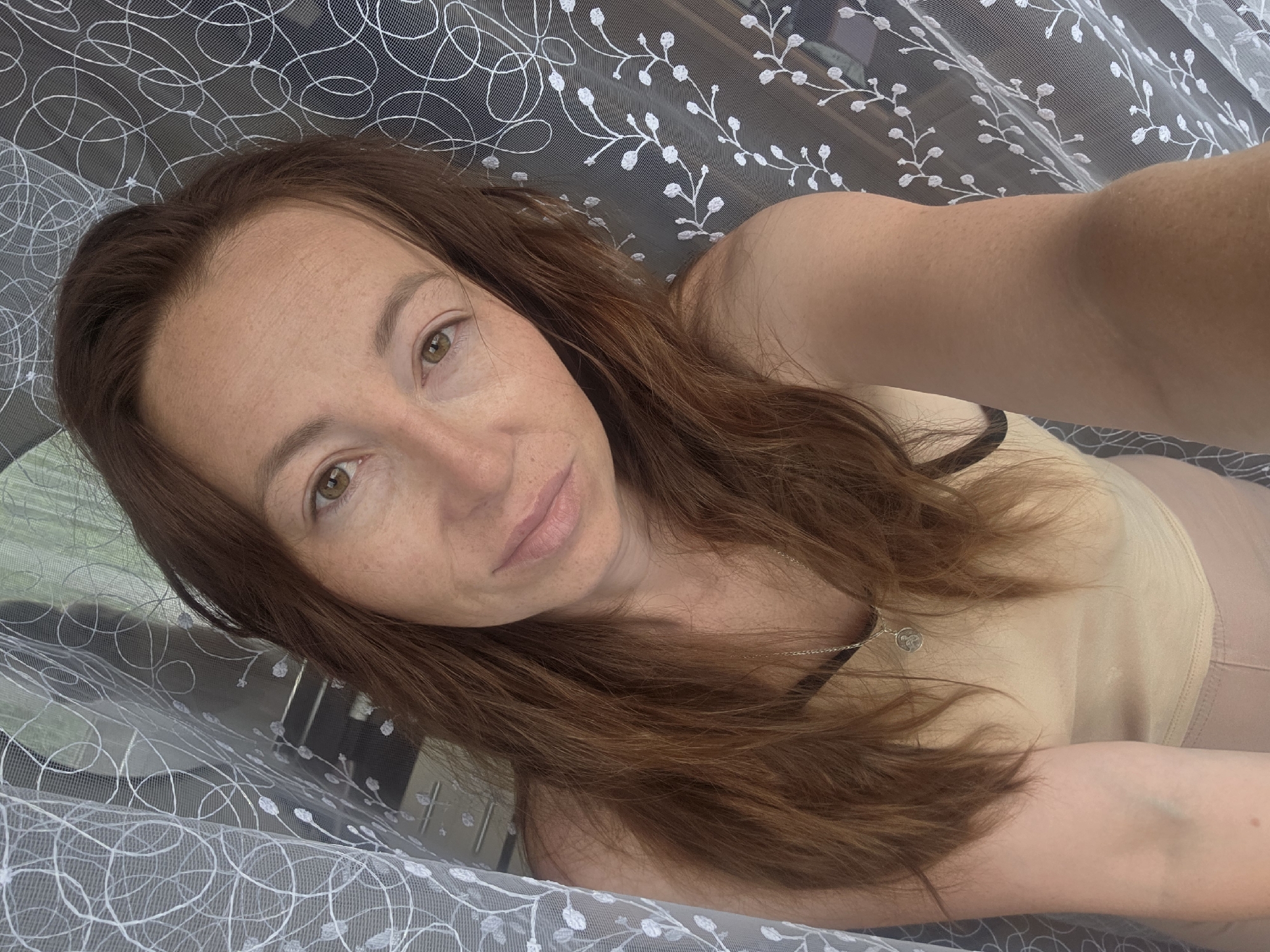 A woman with long, auburn hair and fair skin with freckles is captured in a close-up selfie. She is wearing a light beige tank top and a delicate silver necklace with a circular pendant. She is positioned in front of a white, sheer curtain with a floral pattern. The background is blurred, but hints of a window and possibly outdoor greenery can be discerned. The lighting suggests it is daytime, and the overall ambiance is relaxed and casual. The image appears to have been taken indoors, likely in a residential setting within the city of Chaikovsky, Russia.